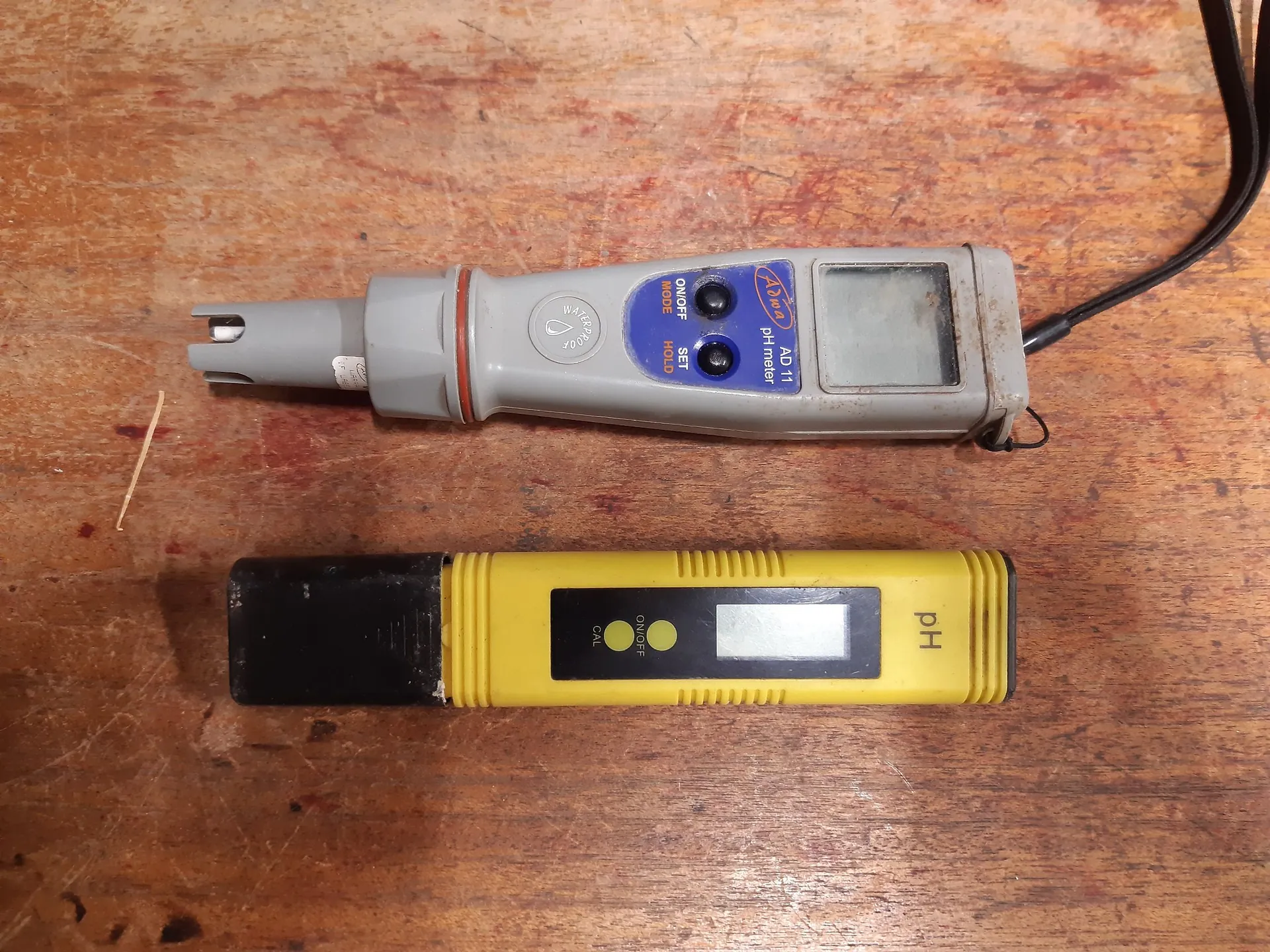 Dwa mierniki pH leżące poziomo na drewnianym blacie. Górne urządzenie jest szare z niebieskimi przyciskami i napisem 'pH meter AD 11'. Dolne urządzenie jest żółte z czarną zakrętką na elektrodę, posiada mały wyświetlacz oraz przyciski 'CAL' i 'ON/OFF', a z prawej strony widnieje napis 'pH'.