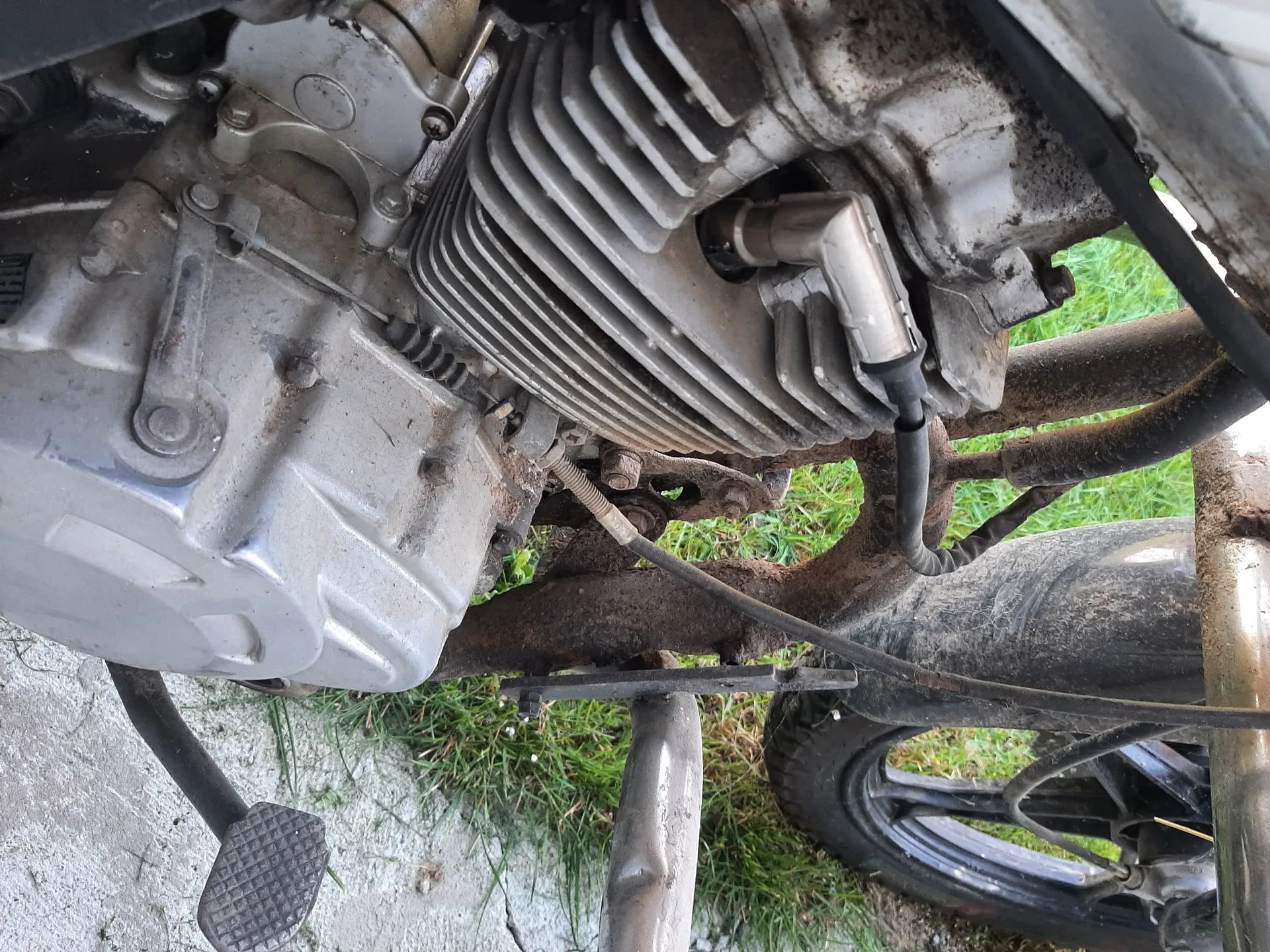 Zbliżenie na silnik motocykla z widocznymi żebrami chłodzenia powietrznego, metalową obudową skrzyni biegów oraz rurą wydechową. W dolnej części widoczny jest podnóżek kierowcy, tylne koło z czarną felgą oraz fragment trawnika.