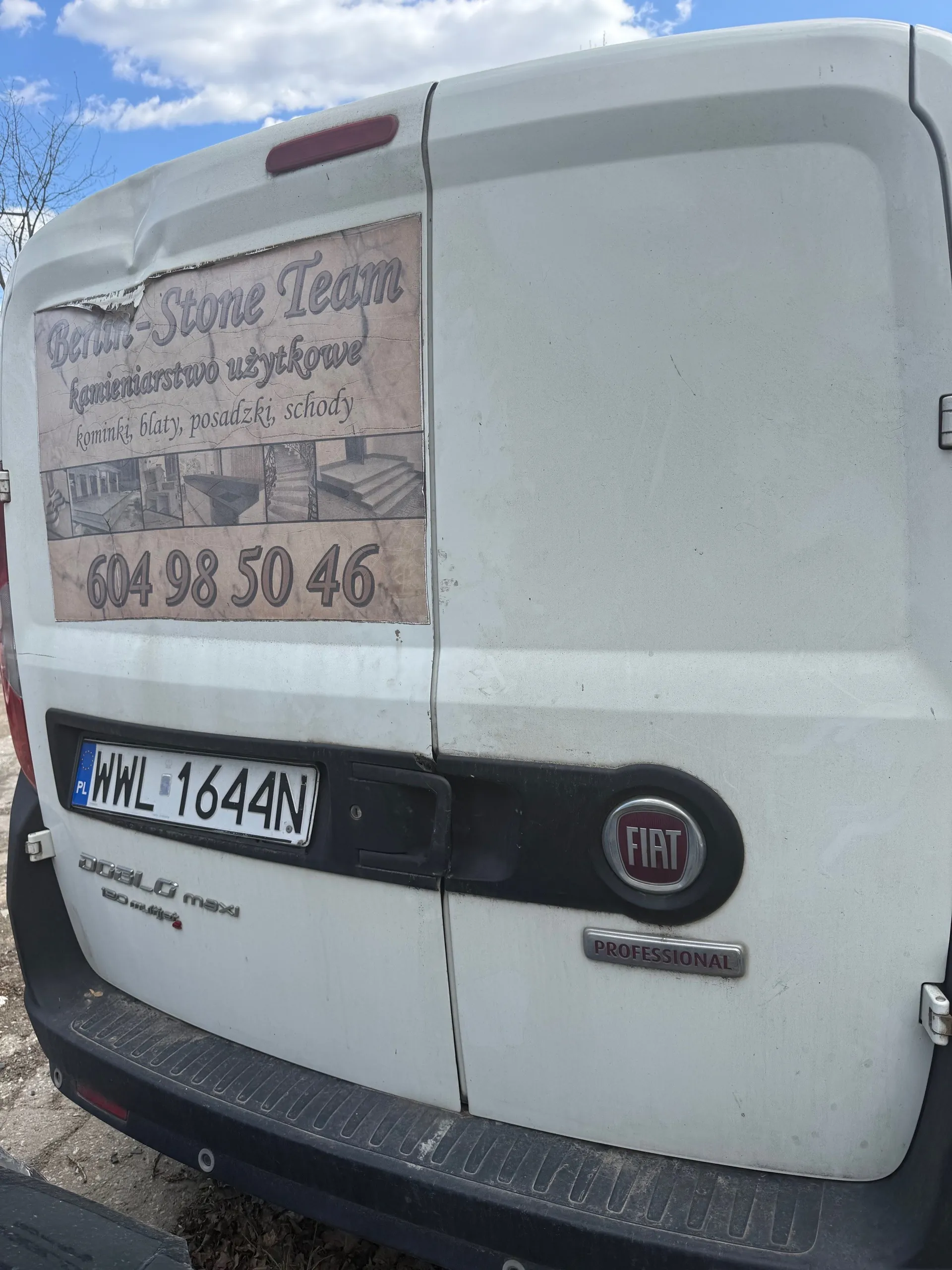 Tył białego furgonetki marki Fiat Professional z widocznym logo FIAT i napisem "PROFESSIONAL". Na lewej stronie tylnych drzwi naklejka z napisem "Berlin-Stone Team", poniżej "kamieniarstwo użytkowe", "kominki, blaty, posadzki, schody" oraz numerem telefonu "604 98 50 46". Tablica rejestracyjna WWL 1644N z kodem kraju PL. Widoczny napis "DOBLO maxi" na dolnej części pojazdu. Tło: niebo z chmurami i drzewo bez liści.