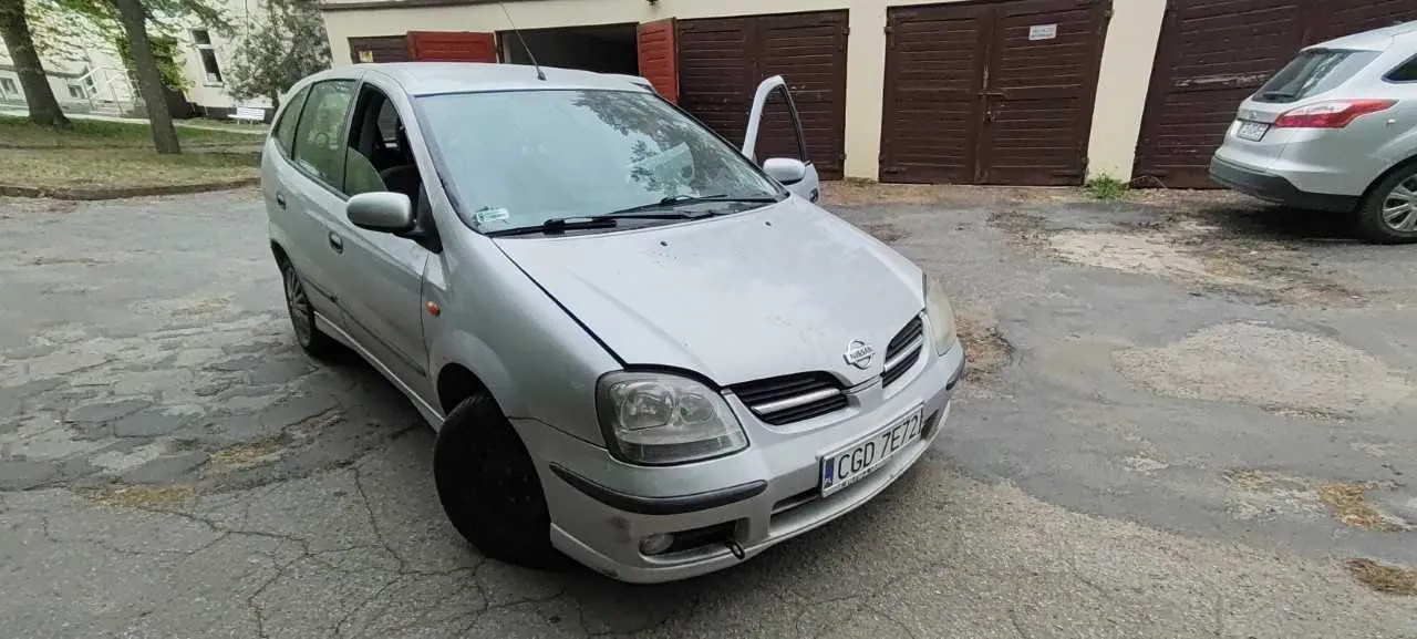 Srebrny samochód marki Nissan z otwartą przednią prawą drzwiami, zaparkowany na popękanej nawierzchni asfaltowej. Widoczny polski numer rejestracyjny CGD 7E72. W tle brązowe garaże, budynek z jasną elewacją oraz drugi srebrny samochód po prawej stronie. Po lewej stronie drzewa i trawnik.