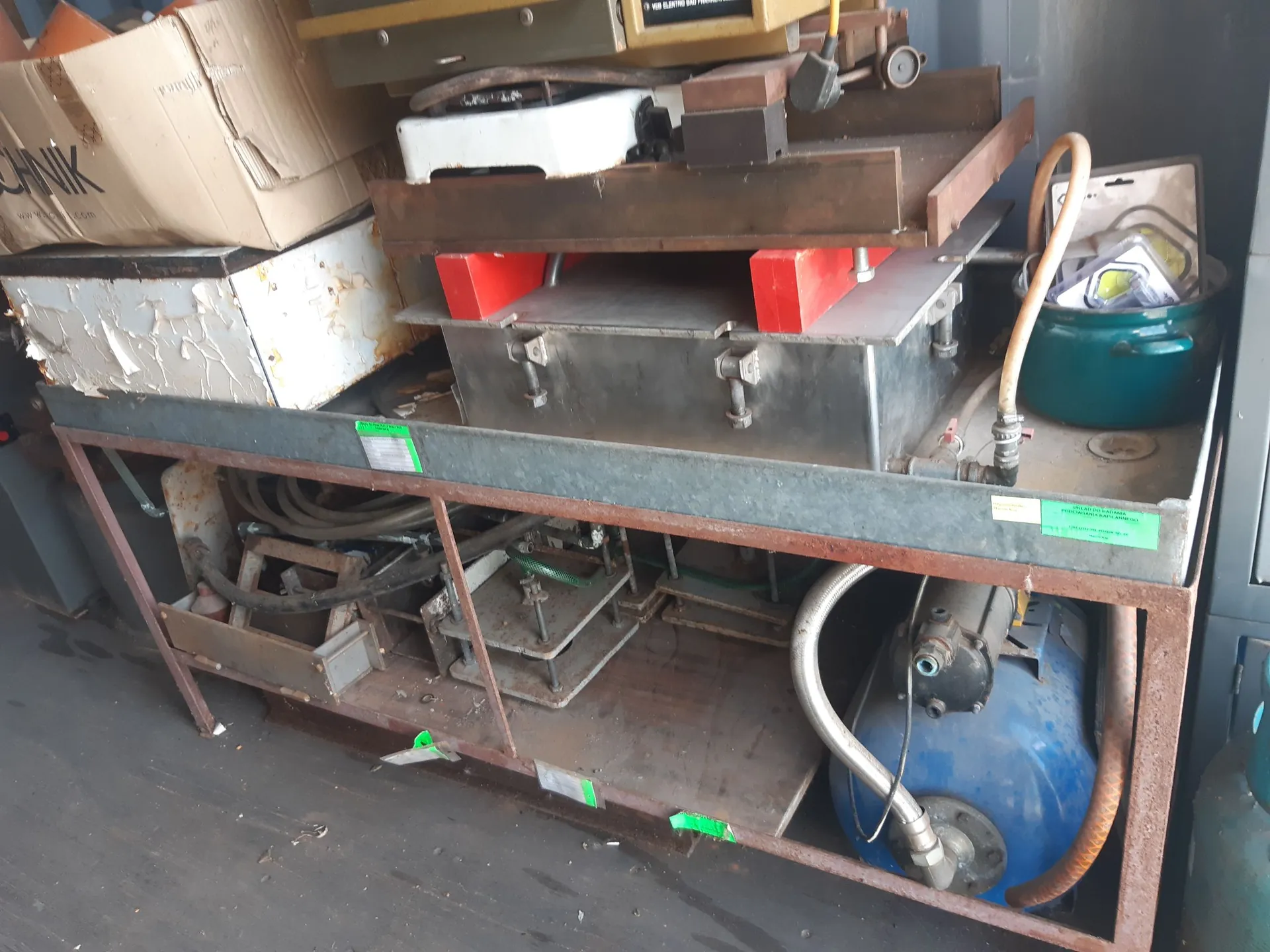 Metalowy regał z dwiema półkami, na których znajduje się różnorodny sprzęt techniczny i warsztatowy. Na górnej półce widoczne są kartonowe pudełka, metalowe formy lub klocki mocujące, czerwone podkładki oraz metalowa kuweta. Po prawej stronie stoi ciemnozielony garnek. Na dolnej półce znajdują się przewody, metalowe elementy konstrukcyjne oraz niebieski zbiornik z pompką i podłączonymi wężami. Na regale przyklejone są małe, zielone etykiety.