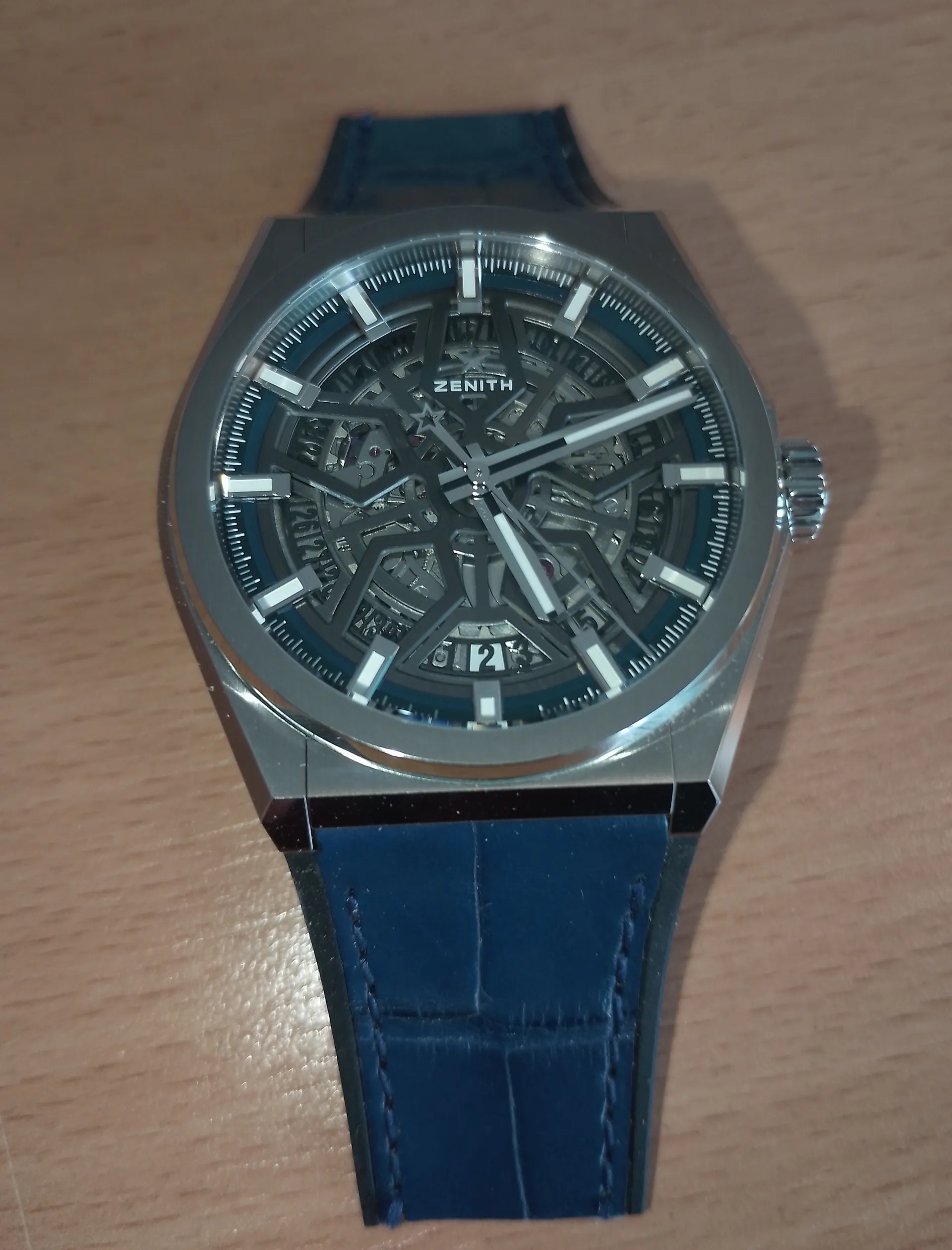 Zegarek marki ZENITH z przezroczystym cyferblatem ukazującym mechanizm. Cyferblat w kolorze niebieskim z srebrnymi indeksami godzinowymi i wskazówkami. Obudowa metalowa, pasek skórzany w kolorze niebieskim z widocznymi szwami. Zegarek umieszczony na jasnobrązowej powierzchni drewnianej. Na cyferblacie widoczny napis ZENITH oraz cyfra 12.