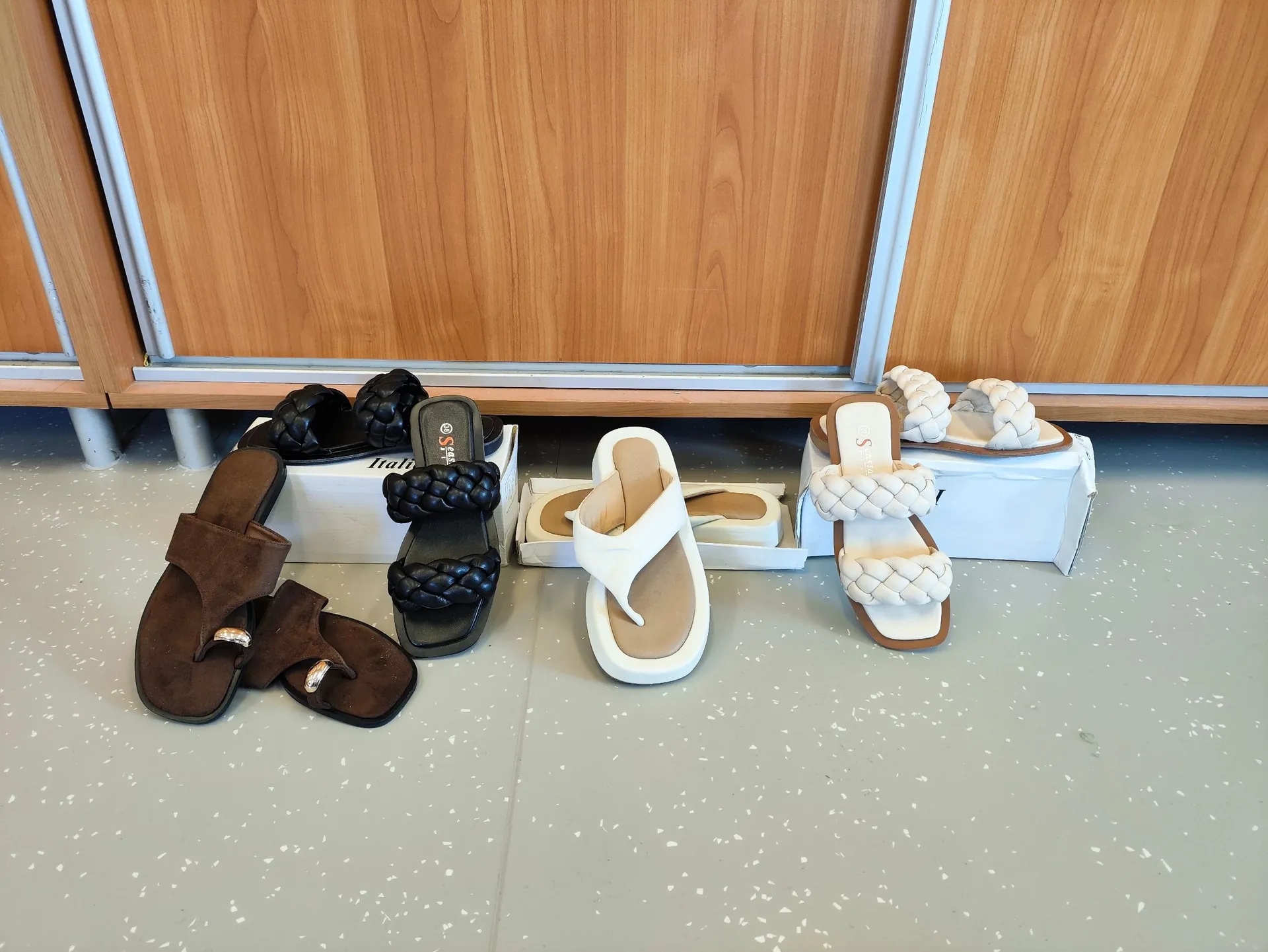 Kilka par damskich sandałów i klapek ułożonych na jasnoszarej podłodze przed drewnianymi szafkami. Od lewej: brązowe zamszowe klapki z dwoma paskami, czarne sandały z grubym plecionym paskiem, białe klapki z jednym paskiem (typu flip-flop) oraz białe sandały z grubym plecionym paskiem. Niektóre buty znajdują się w białych pudełkach.