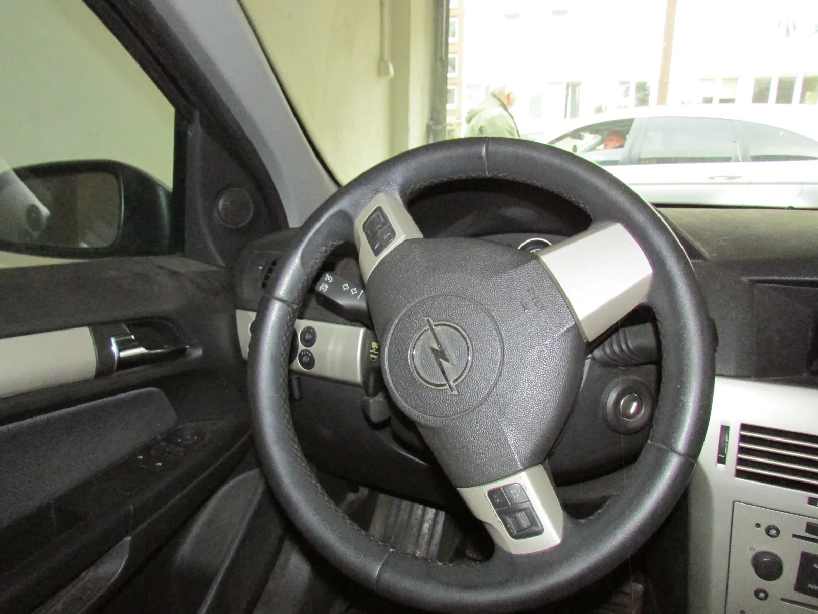 Wnętrze samochodu z widokiem na kierownicę, deskę rozdzielczą i lewe drzwi. Kierownica jest czarna, z logo marki Opel w centralnej części oraz srebrnymi elementami z przyciskami sterującymi. W tle, przez przednią szybę, widoczny jest fragment białego samochodu zaparkowanego na zewnątrz.
