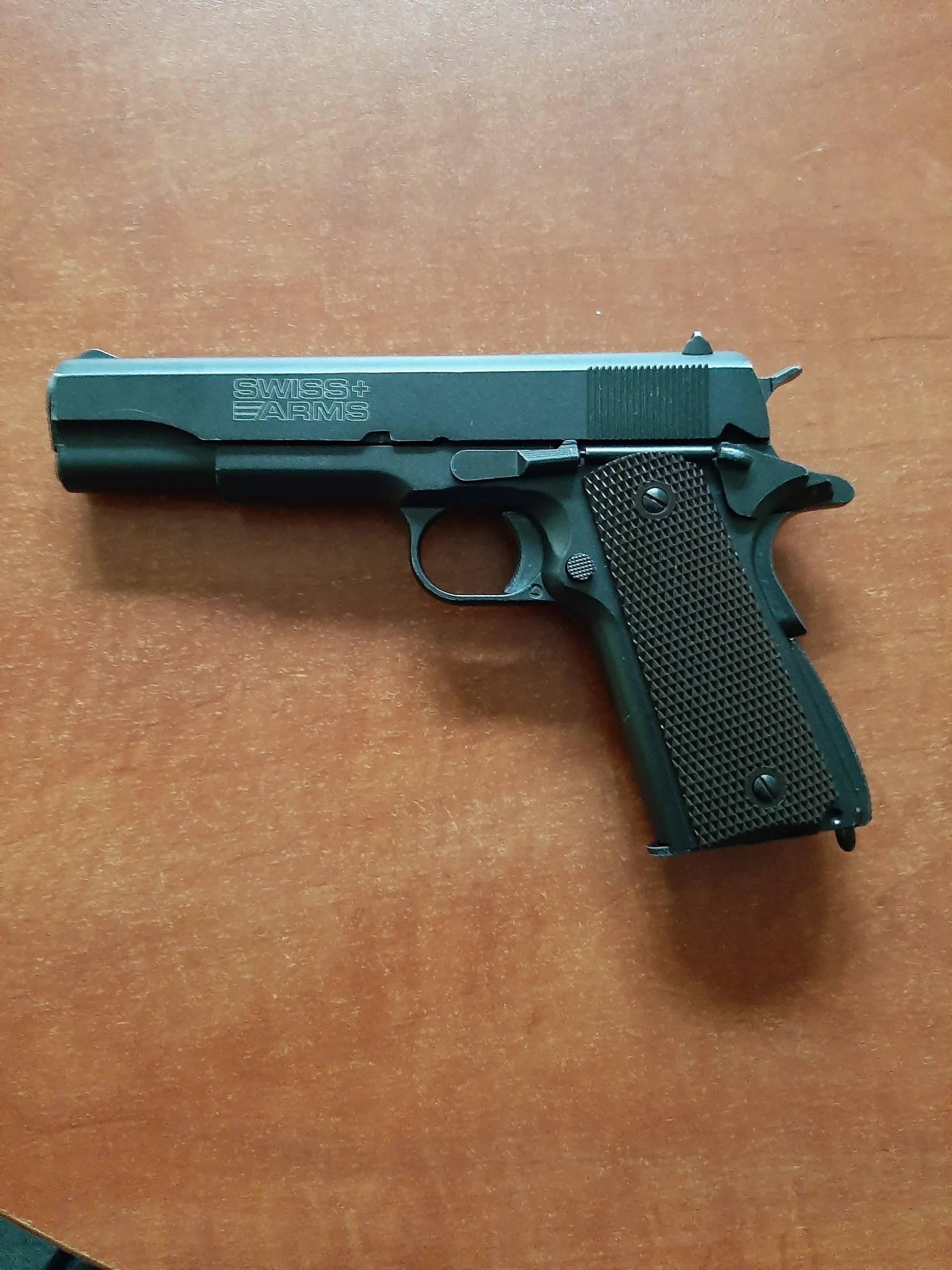 Czarny pistolet marki SWISS+ ARMS leżący pionowo na brązowej powierzchni. Tekst 'SWISS+ ARMS' widoczny na zamku. Uchwyt pistoletu posiada teksturę w kształcie siatki, a na broni znajdują się widoczne śruby i mechanizmy.