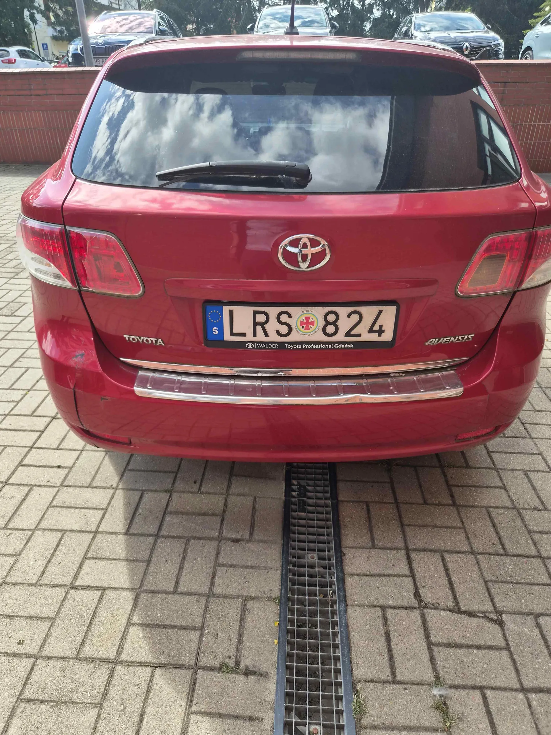 Tylny widok czerwonego samochodu marki Toyota Avensis zaparkowanego na kostce brukowej. Na klapie bagażnika znajduje się tablica rejestracyjna o numerze LRS 824 z oznaczeniem kraju Szwecja (S). Widoczne są napisy 'AVENSIS' oraz 'TOYOTA'.