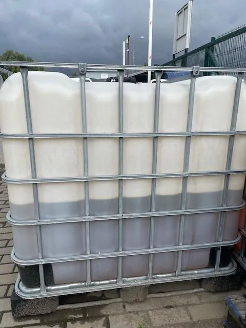 Biały plastikowy zbiornik IBC w metalowej klatce ochronnej, wypełniony w połowie szarą cieczą. Zbiornik stoi na betonowym podłożu pod pochmurnym niebem.