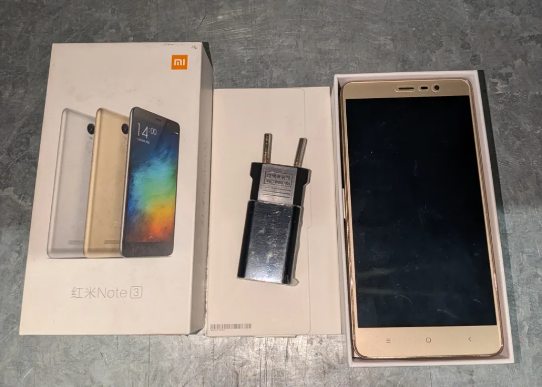 Zestaw produktów marki Xiaomi ułożony na szarej, teksturowanej powierzchni. Po lewej stronie znajduje się białe pudełko z logo Xiaomi i napisem 'Note 3', na którym widnieją grafiki trzech smartfonów w kolorach srebrnym, złotym i czarnym. W środku znajduje się białe pudełko z czarną ładowarką sieciową z wtyczką typu C. Po prawej stronie, w białym opakowaniu, leży złoty smartfon z wyłączonym, czarnym ekranem i widocznymi przyciskami nawigacyjnymi na dolnej ramce.