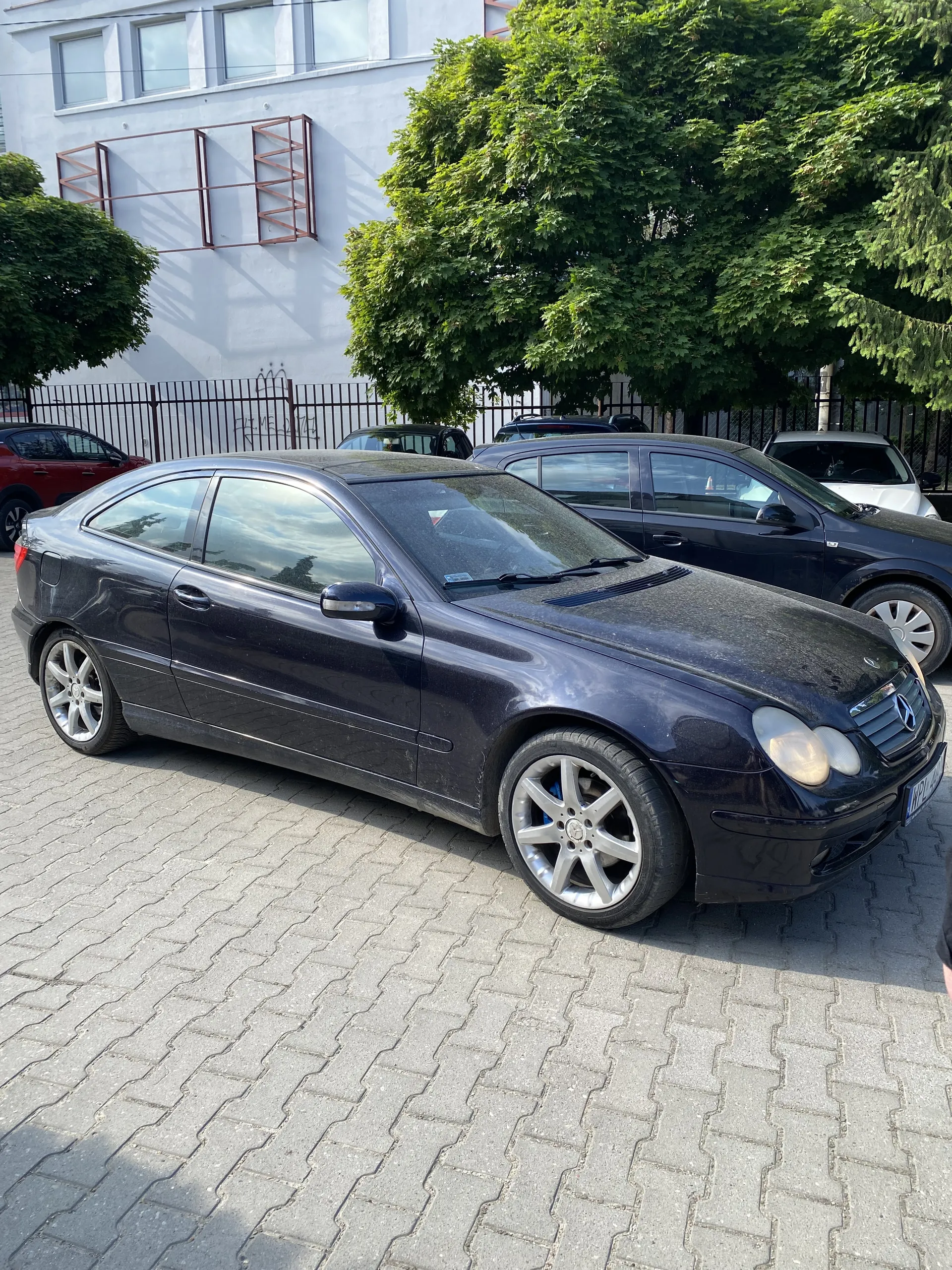 Trzy samochody zaparkowane na brukowanej powierzchni. Na pierwszym planie czarny samochód osobowy z srebrnymi felgami. Za nim czarny i czerwony samochód. Po lewej stronie zielone drzewa i budynek z białą fasadą i oknami. Widoczne metalowe ogrodzenie.