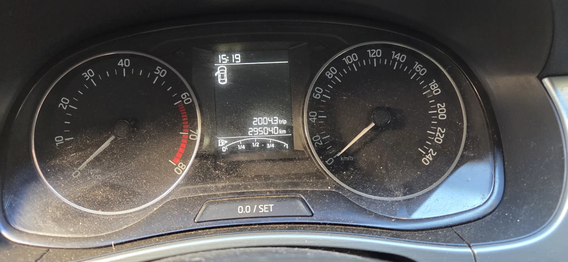 Zbliżenie na deski rozdzielcze samochodu. Po lewej stronie wskaźnik prędkości z zakresem 0-240 km/h, po prawej obrotomierz z zakresem 0-8000 obr./min (oznaczony jako x1000 rpm). W centralnym wyświetlaczu cyfrowym widoczne informacje: '20043 trip', '295040 km', godzina '15:19' oraz ikona samochodu z otwartą klapą bagażnika. Na górze deski napis '0.0 / SET'. Całość pokryta jest kurzem. Wskaźniki i tekst na wyświetlaczu są odwrócone do góry nogami.