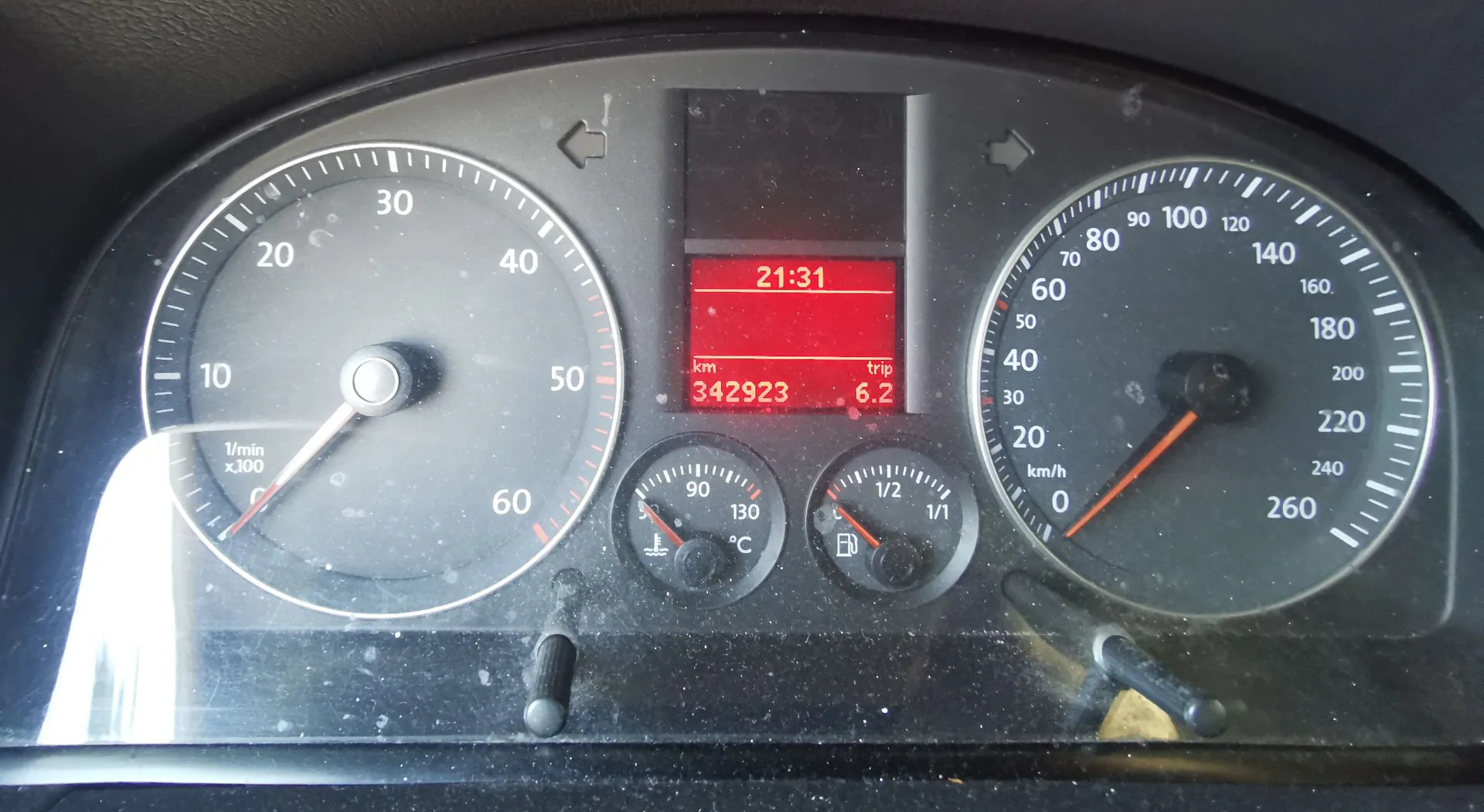 Zbliżenie na deskę rozdzielczą samochodu. Po lewej stronie znajduje się obrotomierz z zaznaczonymi wartościami od 10 do 60 (x100 1/min). W centrum znajduje się czerwony wyświetlacz cyfrowy pokazujący godzinę 21:31, całkowity przebieg 342923 km oraz licznik trip 6.2. Poniżej wyświetlacza są dwa mniejsze wskaźniki: temperatura cieczy chłodzącej oraz poziom paliwa. Po prawej stronie znajduje się prędkościomierz z zaznaczonymi wartościami od 0 do 260 km/h.