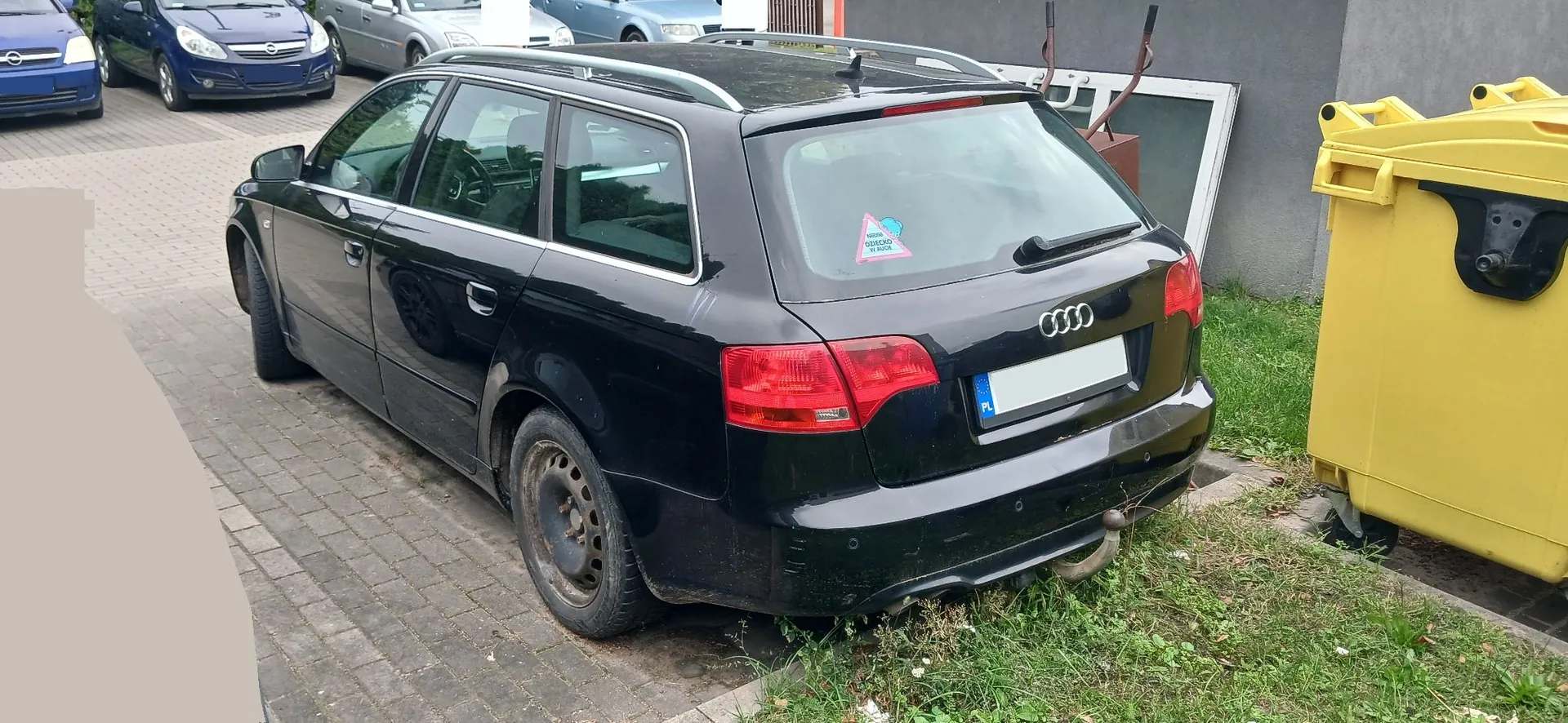 Czarny samochód marki Audi w nadwoziu kombi zaparkowany na kostce brukowej. Pojazd jest widoczny od tyłu i z boku. Na tylnej szybie znajduje się mała, kolorowa naklejka. Po prawej stronie obrazu stoi duży, żółty kontener na odpady. W tle widoczne są inne zaparkowane samochody oraz fragment szarego budynku.