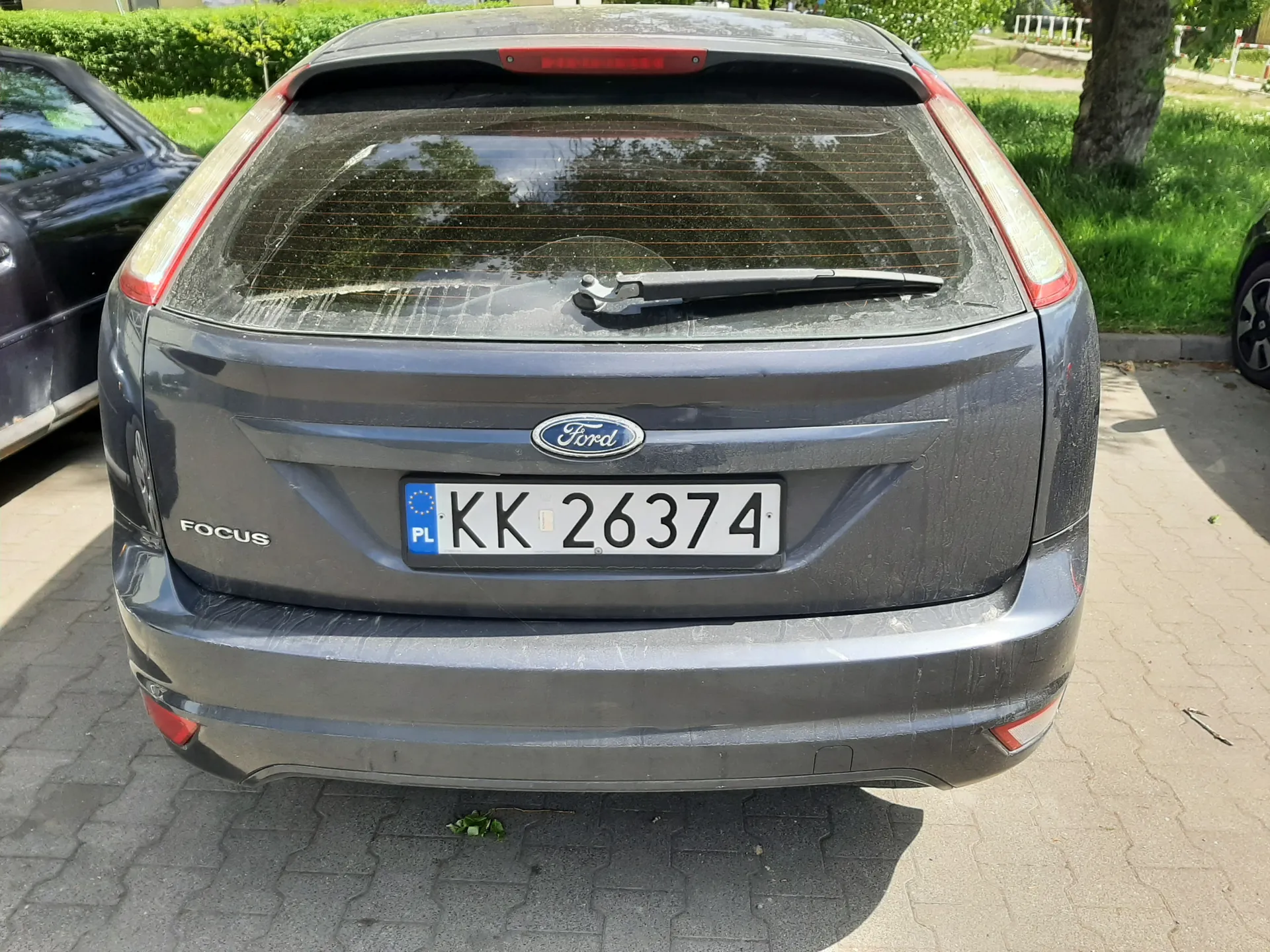 Tył szarego samochodu Ford Focus z tablicą rejestracyjną KK 26374 (PL) na tle zielonej trawy, krzewów i drzewa. Widoczne są również częściowo inne zaparkowane pojazdy oraz brukowana powierzchnia. Na bagażniku umieszczono logo Ford oraz napis FOCUS po lewej stronie.
