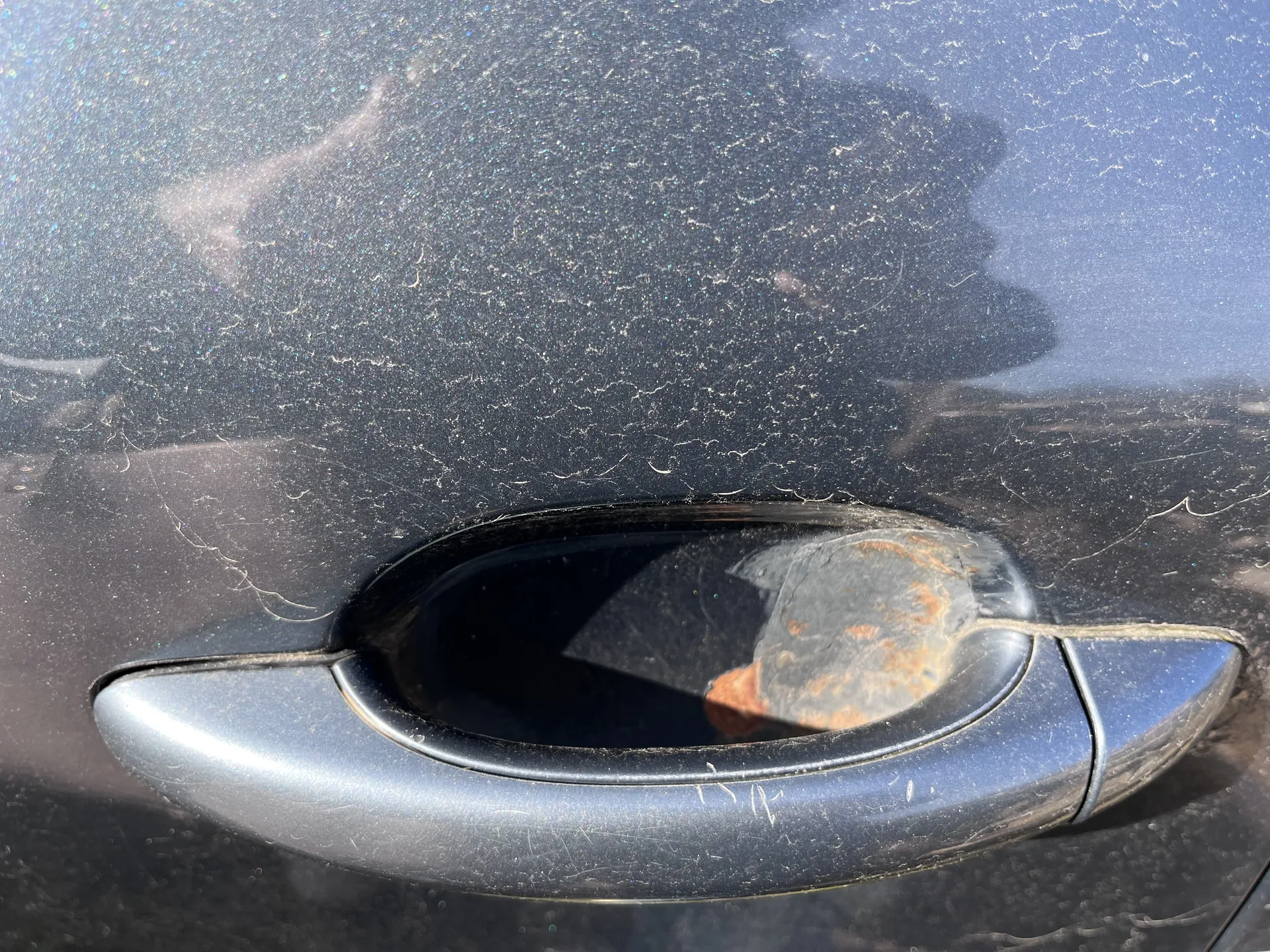 Zbliżenie na metalową klamkę drzwi samochodu w kolorze ciemnoszarym lub czarnym. Powierzchnia drzwi pokryta jest licznymi drobnymi zadrapaniami i kurzem. W klamce odbija się sylwetka osoby trzymającej aparat lub telefon, z widocznymi pomarańczowymi plamami na odbiciu. Tło jest rozmyte, ale widać fragmenty otoczenia odbijające się w powierzchni samochodu.