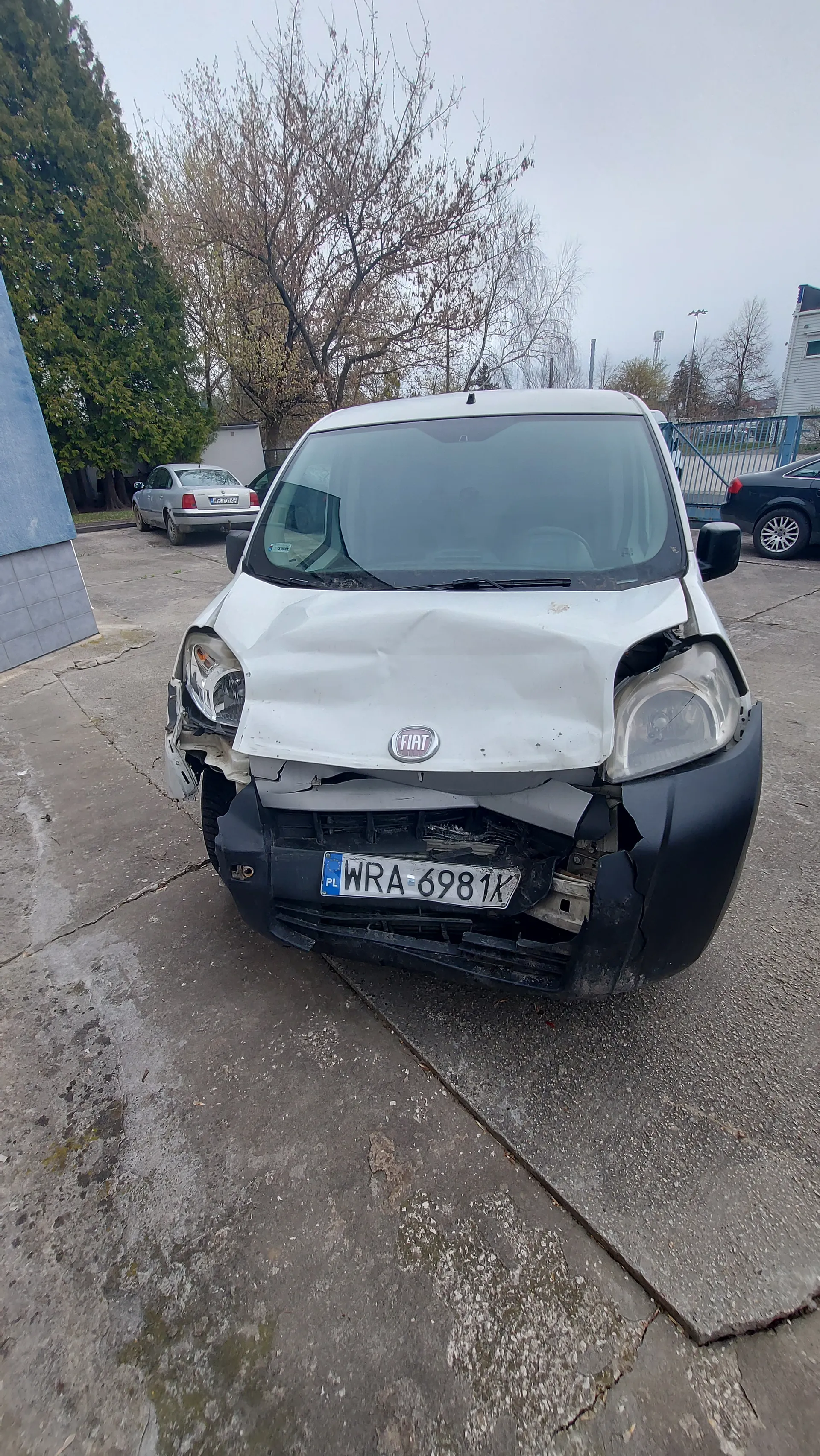 Biały samochód marki Fiat z uszkodzonym przodem, widoczna tablica rejestracyjna WRA 6981K. W tle inne pojazdy, drzewa i budynki. Niebo jest pochmurne.