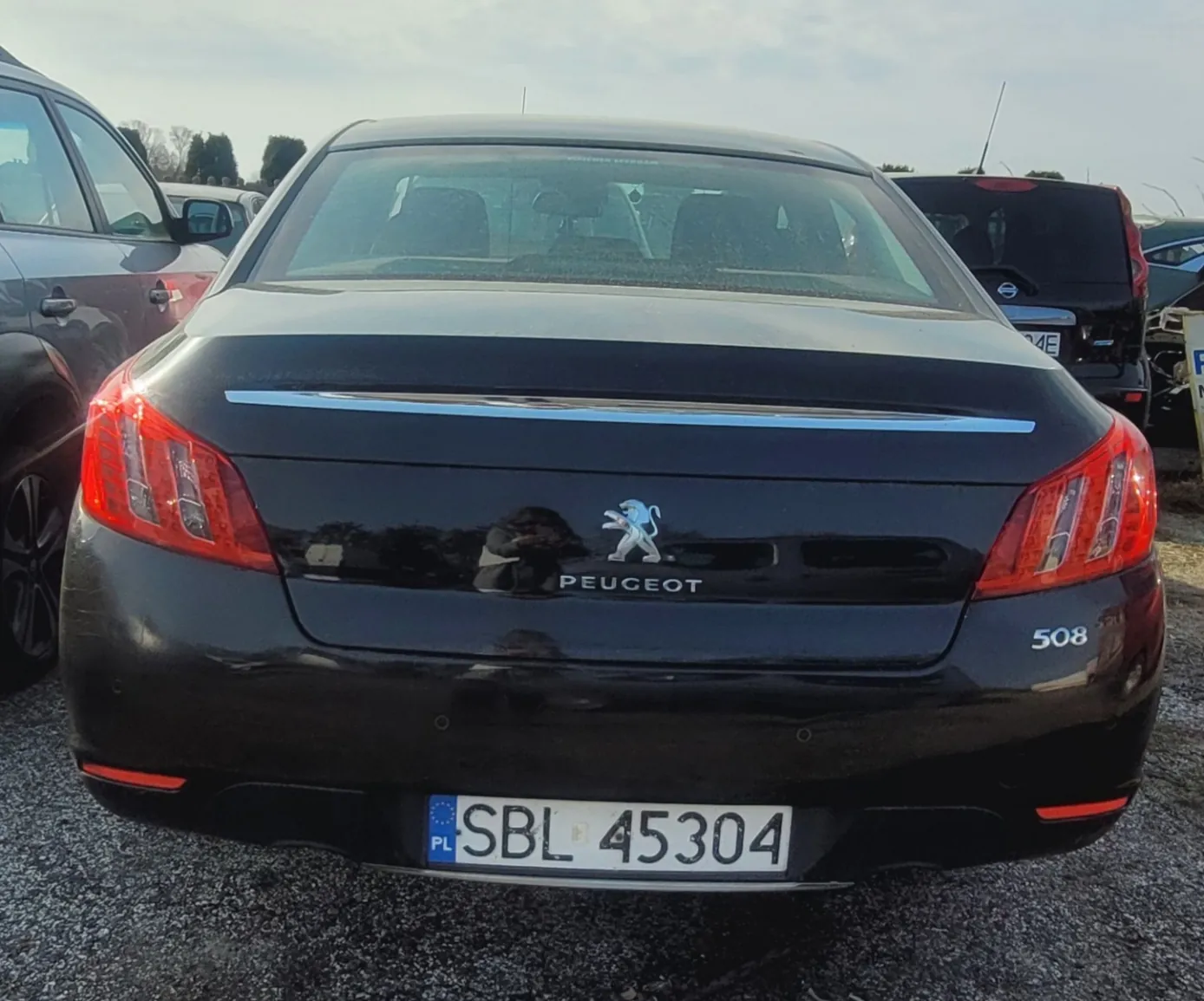 Tył czarnego samochodu marki Peugeot model 508 z widocznym logo i napisem "PEUGEOT" na bagażniku. Tablica rejestracyjna z polskim kodem PL i numerem SBL 45304. Samochód zaparkowany na żwirowym placu parkingowym, w tle inne pojazdy oraz częściowo zachmurzone niebo.