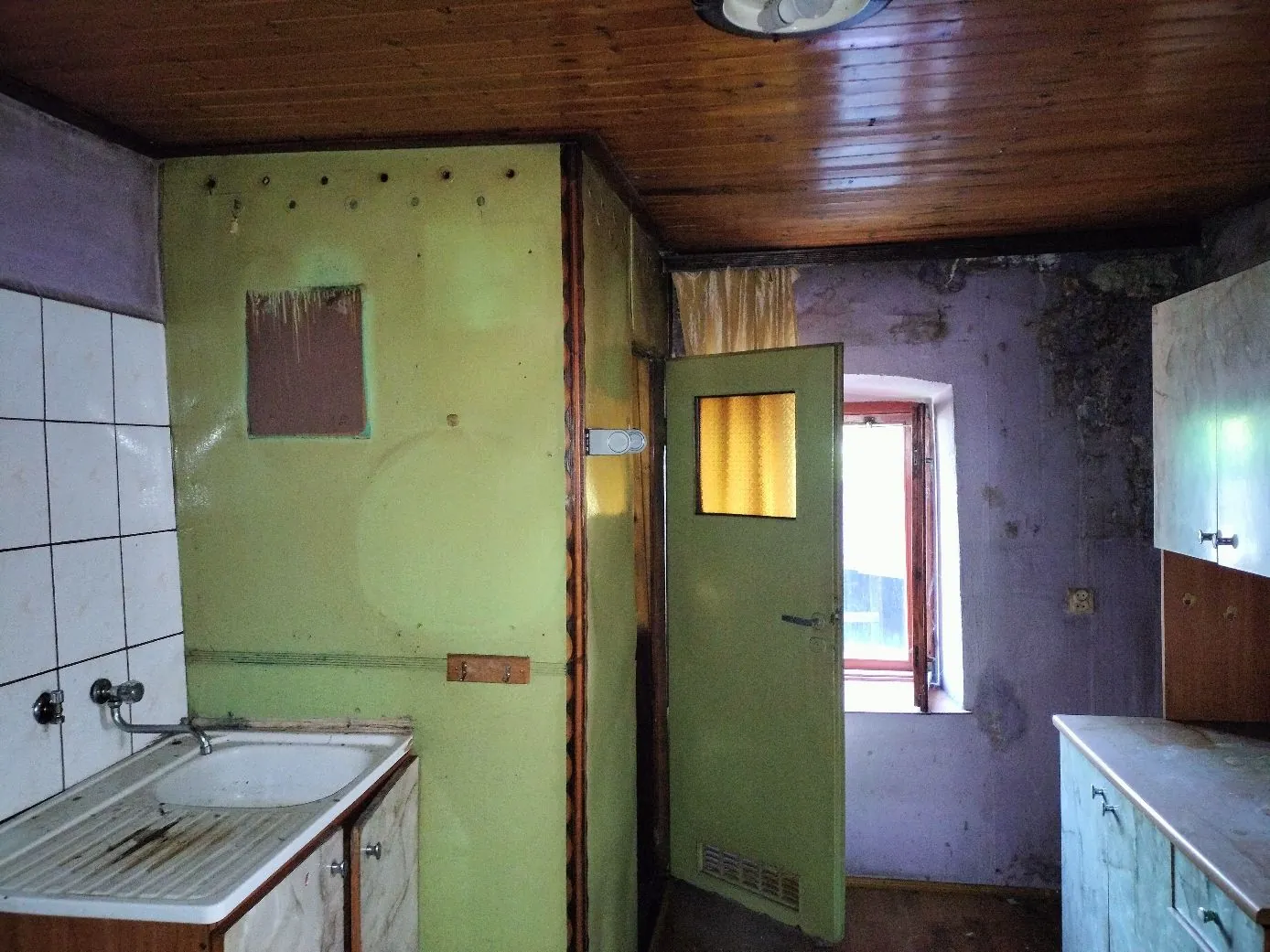 Wnętrze zaniedbanego pomieszczenia, prawdopodobnie łazienki lub kuchni. Po lewej stronie znajduje się biały zlew wbudowany w szafkę, nad nim białe kafelki na ścianie. Ściana w centrum jest pomalowana na jasnozielony kolor i jest w złym stanie, z widocznym prostokątnym ubytkiem w tynku. Po prawej stronie znajduje się otwarte zielone drzwi z małym, kwadratowym okienkiem, prowadzące do innego pomieszczenia z widocznym oknem. Sufit jest wykonany z ciemnych drewnianych desek. Ściany po prawej stronie są w kolorze fioletowym, z widocznymi śladami wilgoci i odpadającego tynku.
