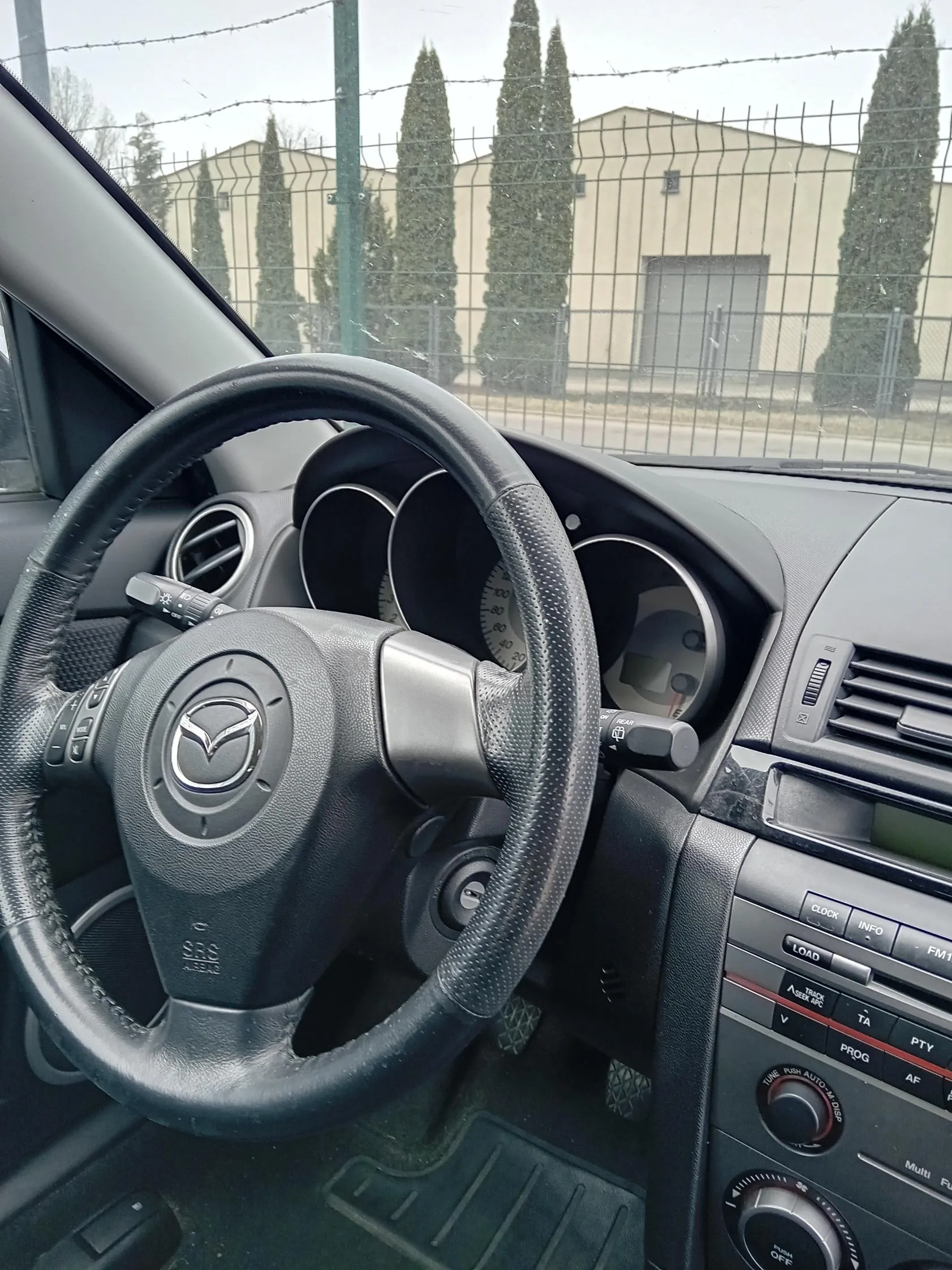 Wnętrze samochodu marki Mazda z widocznym kierownicą z logo producenta, przyciskami sterującymi po lewej stronie kierownicy oraz tablicą przyrządów z okrągłymi wskaźnikami. W centralnej konsoli widoczne są przyciski oznaczone napisami: CLOCK, INFO, LOAD, FM1, TA, PTY, PROG, AF oraz pokrętła regulacyjne. Za przednią szybą widoczny jest ogrodzenie z drutem kolczastym, wysokie drzewa i budynek przemysłowy. Niebo jest pochmurne.