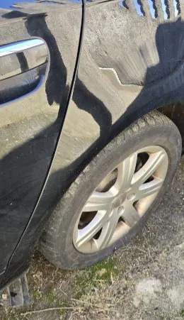 Czarny samochód widziany z boku, z bliska przednie koło z srebrnym felgiem wieloramiennym. Opona jest brudna, a na ziemi widoczna trawa i kurz. Fragment drzwi z uchwytem oraz cień na karoserii.