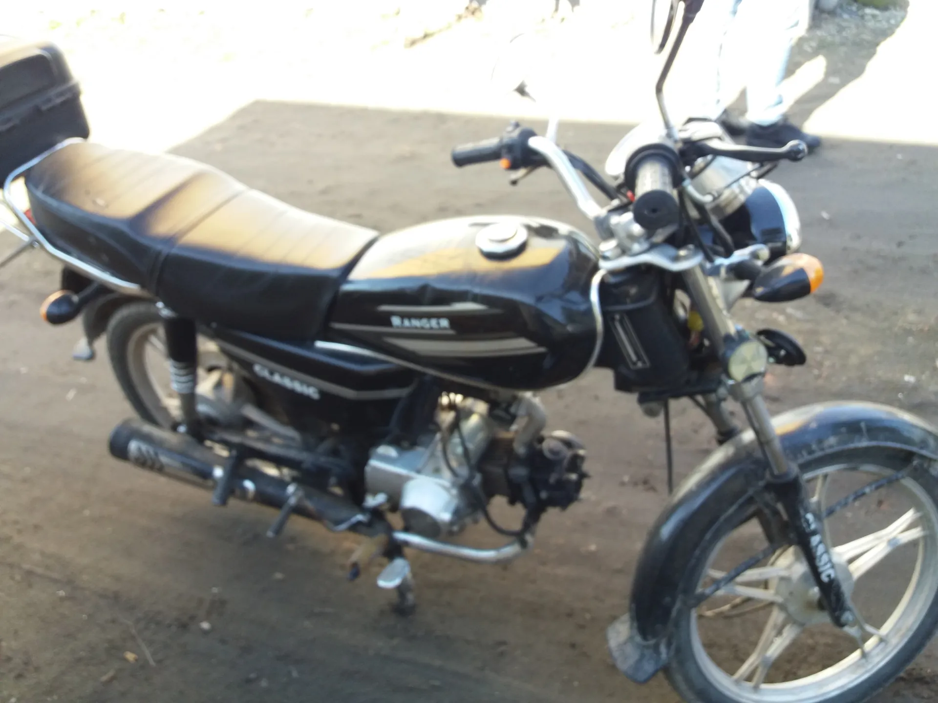 Czarny motocykl marki Ranger z napisem Classic na boku. Widoczne są czarne siedzenie, bagażnik na tylnej części, silnik oraz przednie koło. Na zbiorniku paliwa znajduje się napis Ranger, a na ramie i przednim błotniku napis Classic. W tle widoczna osoba w jasnych spodniach i czarnych butach stojąca na asfaltowej powierzchni.