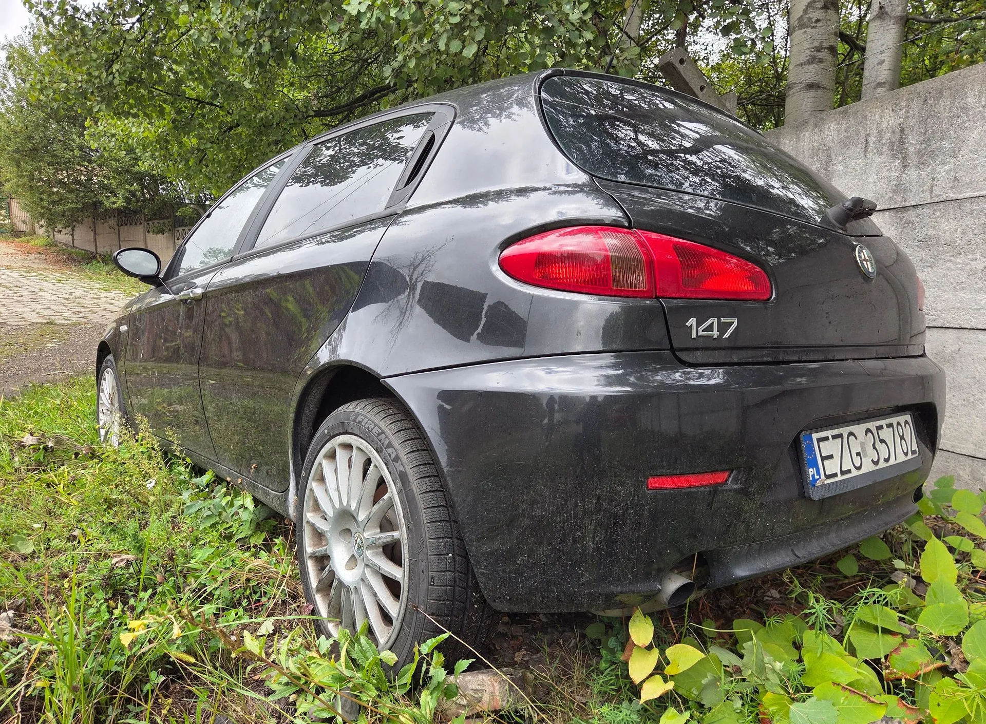 Czarny samochód Alfa Romeo 147 zaparkowany na trawiastym poboczu obok betonowego muru. Widoczny jest tył i bok pojazdu, tylna lampa, oznaczenie modelu '147' oraz tablica rejestracyjna o numerze EZG 35782. Samochód jest zakurzony i otoczony zieloną roślinnością.