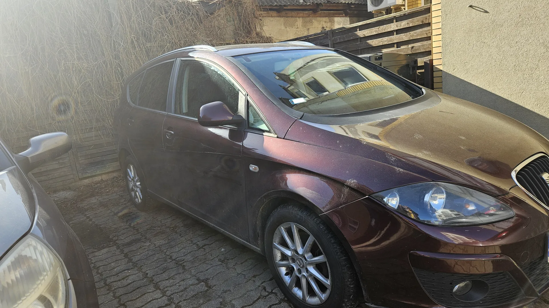 Ciemnoczerwony samochód typu kombi zaparkowany na brukowanej powierzchni. Po lewej stronie widoczna część innego samochodu. W tle drewniane ogrodzenie, suche pnącza, betonowa ściana i budynek z drewnianą konstrukcją. Słoneczne oświetlenie z lekkim efektem flary.