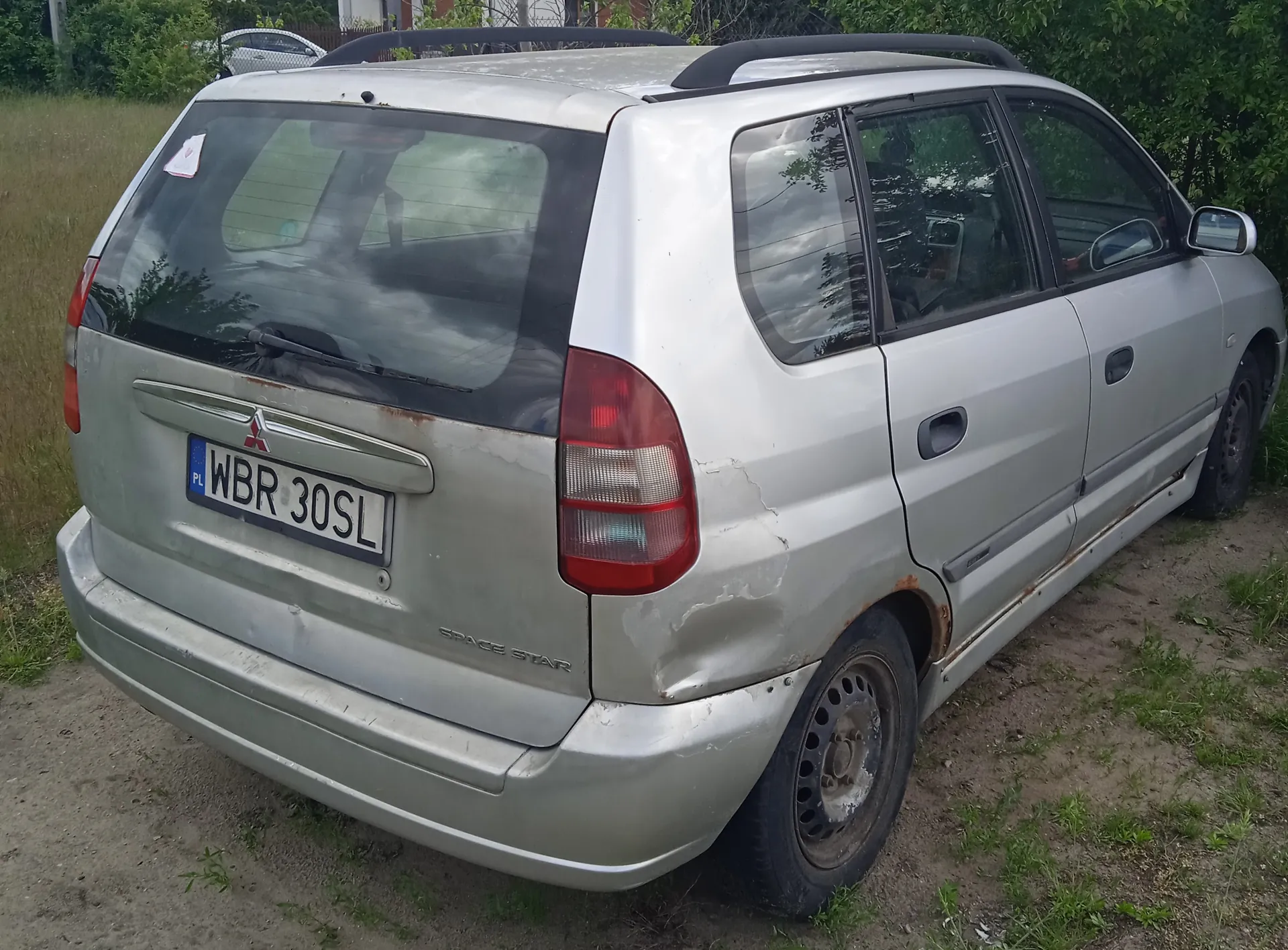 Srebrny samochód Mitsubishi Space Star zaparkowany na terenie z trawą i ziemią. Widok z tyłu i boku. Na tablicy rejestracyjnej widoczny napis WBR 30SL oraz oznaczenie PL. Samochód posiada dachówkę, zarysowane ślady rdzy na karoserii, zwłaszcza w okolicach tylnego koła. W tle zielone krzewy, drzewa oraz inny samochód za ogrodzeniem. Na tylnej szybie naklejka.