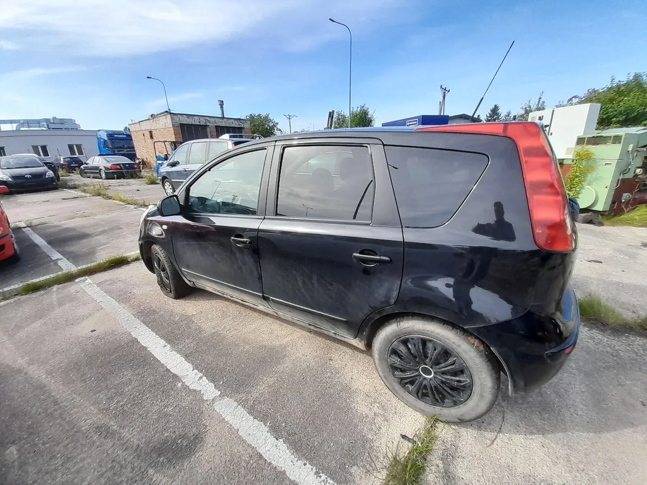 Czarny samochód typu minivan zaparkowany na betonowym placu. Pojazd jest pokazany z profilu od strony pasażera. W tle widoczne są inne samochody, w tym biały van i niebieska ciężarówka, a także niskie budynki przemysłowe pod jasnoniebieskim niebem z lekkimi chmurami.