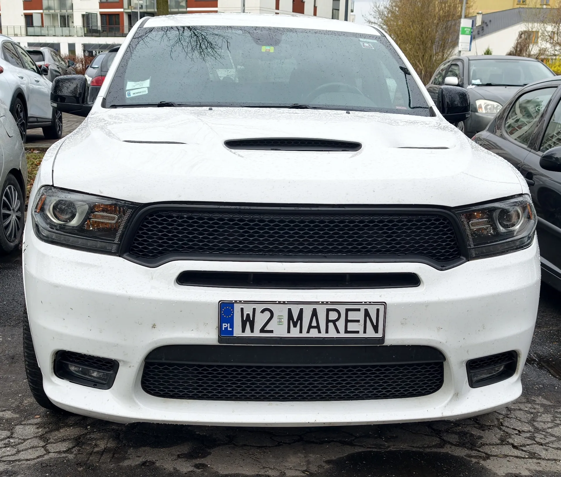 Biały samochód z przodu, z widoczną tablicą rejestracyjną PL W2 MAREN. Samochód ma czarną kratę chłodnicy, reflektory i krople wody na maskie. W tle inne zaparkowane pojazdy, budynki mieszkalne oraz mokry asfalt.