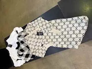 Biały szalik z czarnym wzorem powtarzających się liter G, czarna etykieta z napisem GUCCI, biała etykieta oraz czarno-biały szachownicowy przedmiot leżące na ciemnej powierzchni. Tło stanowi jasna podłoga.