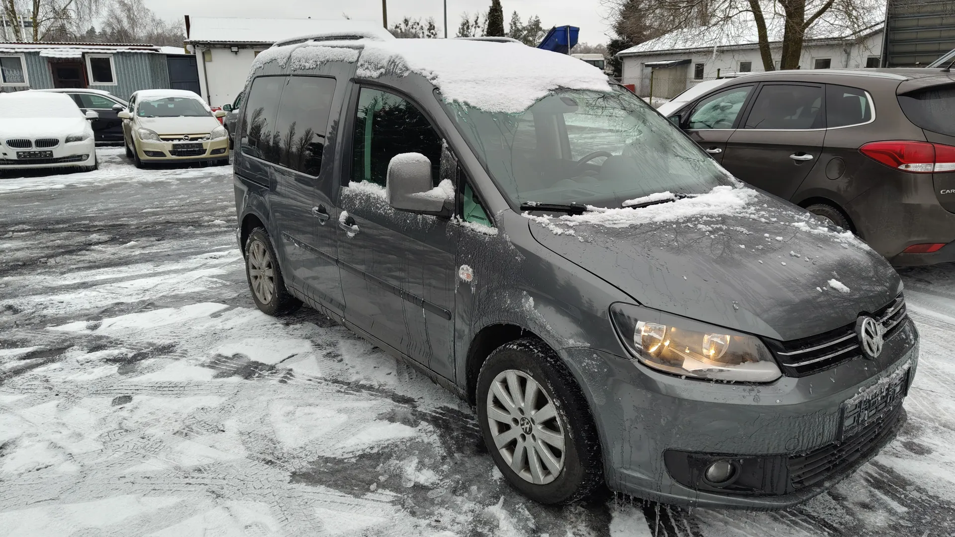 Szary samochód osobowy marki Volkswagen z pokrytą śniegiem maską i dachem, zaparkowany na śnieżnym parkingu. W tle widoczne są inne pojazdy: biały samochód BMW, beżowy Opel oraz brązowy samochód osobowy. Tło stanowią budynki z dachami pokrytymi śniegiem, drzewa bez liści i szare niebo. Na masce i dachu pojazdu leży warstwa śniegu, a z przodu wiszą krople lodu. Droga pokryta jest śniegiem i lodem z widocznymi śladami opon.