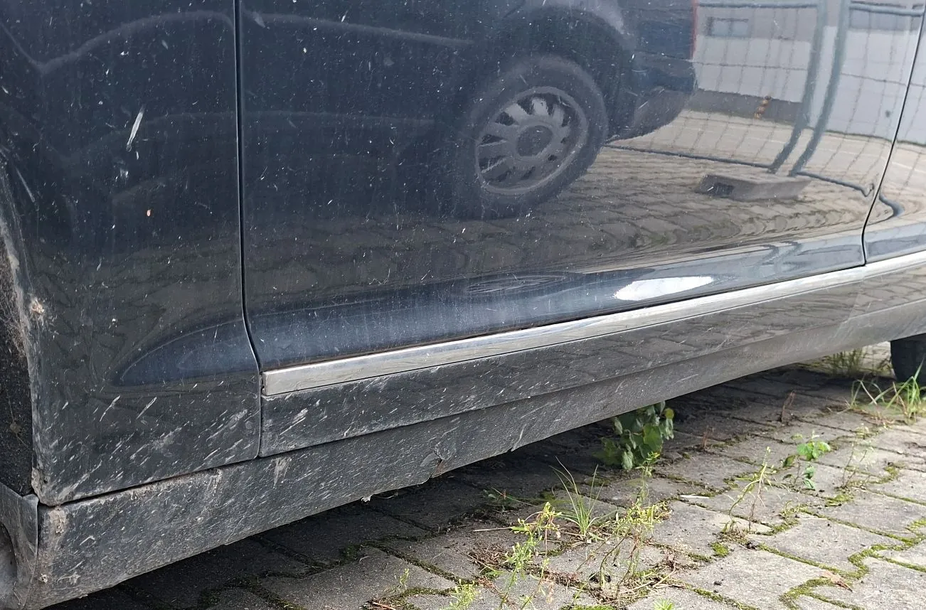 Zbliżenie na dolną część boku ciemnego samochodu. Widoczny jest próg auta z chromowaną listwą, który jest zabrudzony pyłem i błotem. Samochód stoi na kostce brukowej, z której wyrastają kępki trawy i małe rośliny.