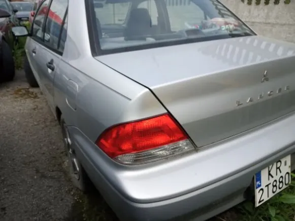 Srebrny samochód Mitsubishi Lancer widziany od tyłu, zaparkowany na żwirowej powierzchni. Widoczny czerwony tylny reflektor, tablica rejestracyjna z napisem KR 2T880 oraz logo marki i nazwa modelu Lancer na bagażniku. W tle fragment innych pojazdów i kamienna ściana.