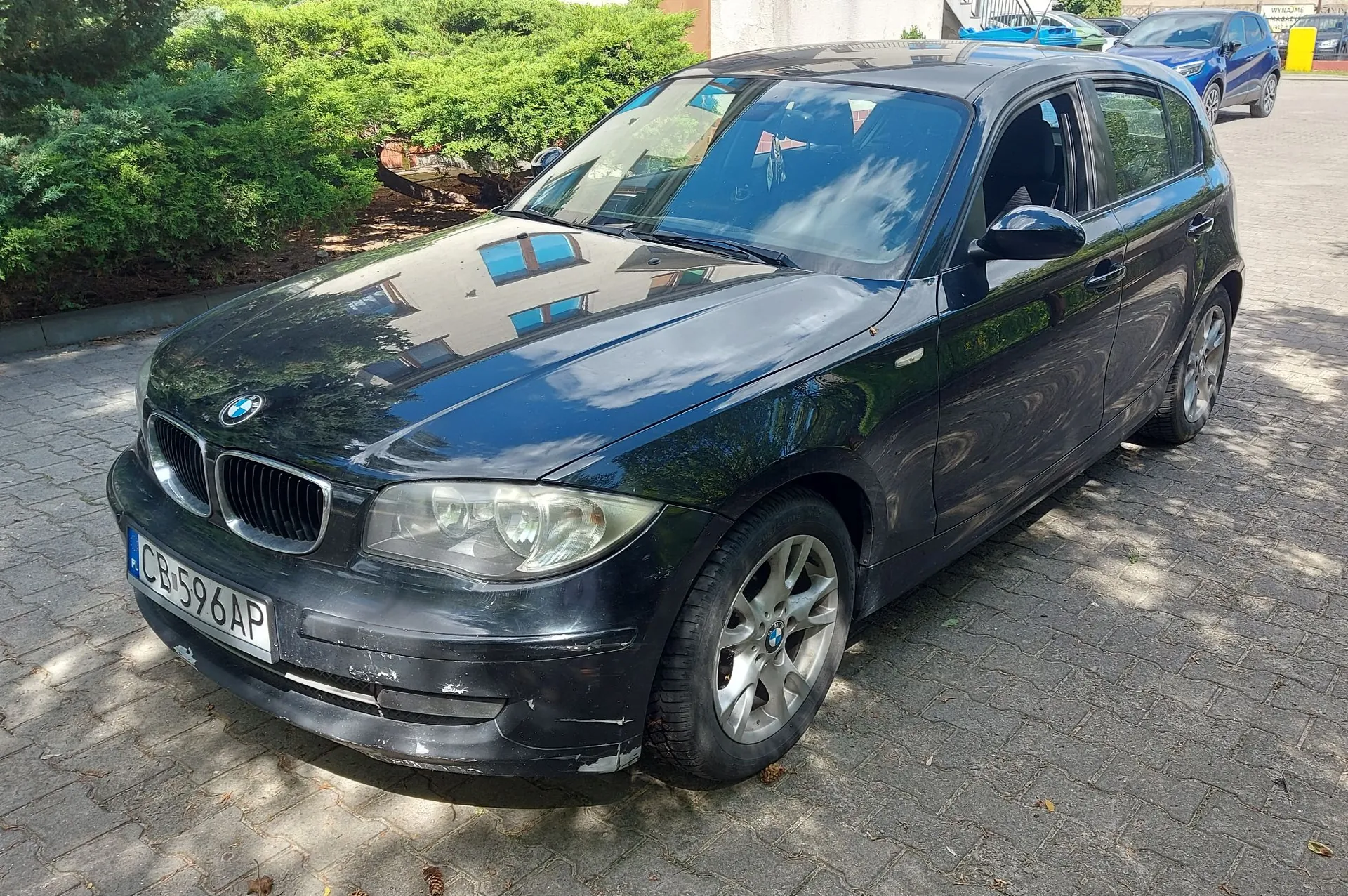 Czarny samochód marki BMW zaparkowany na kostce brukowej. Pojazd jest widoczny z przodu i z boku. Na przednim zderzaku widoczne są zarysowania i uszkodzenia lakieru. Tablica rejestracyjna ma numer CB 596AP. W tle widać zielone krzewy oraz inne zaparkowane samochody.