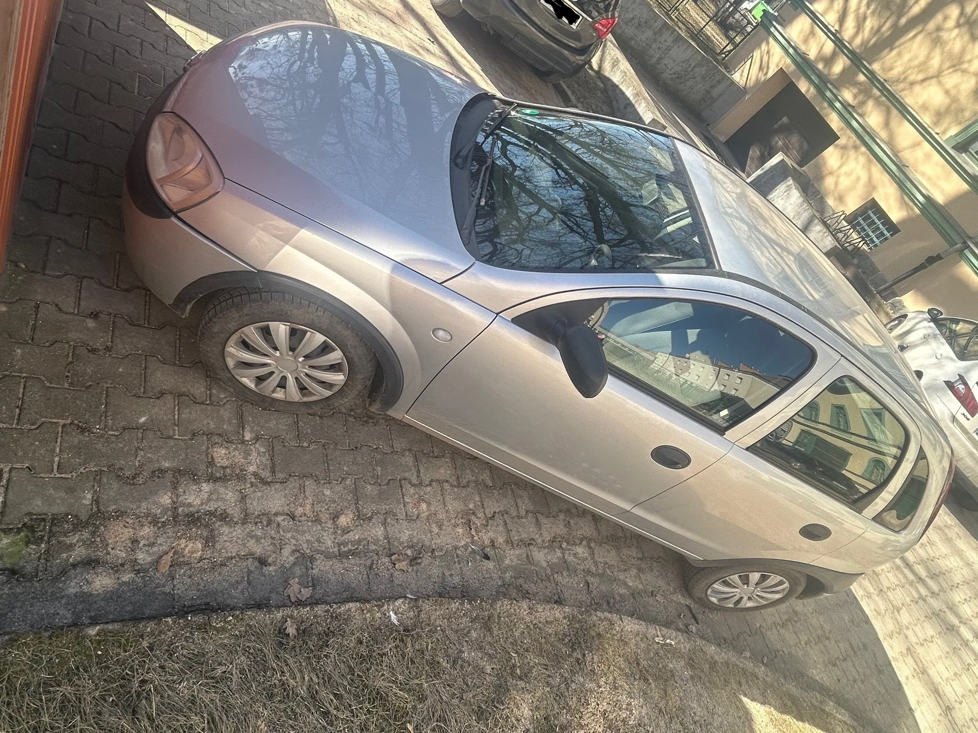 Srebrny samochód osobowy typu hatchback zaparkowany na brukowanej powierzchni. W tle widoczny budynek z zielonymi rurami, inne zaparkowane samochody oraz sucha trawa po prawej stronie. Przez szyby odbijają się budynki i drzewa bez liści.