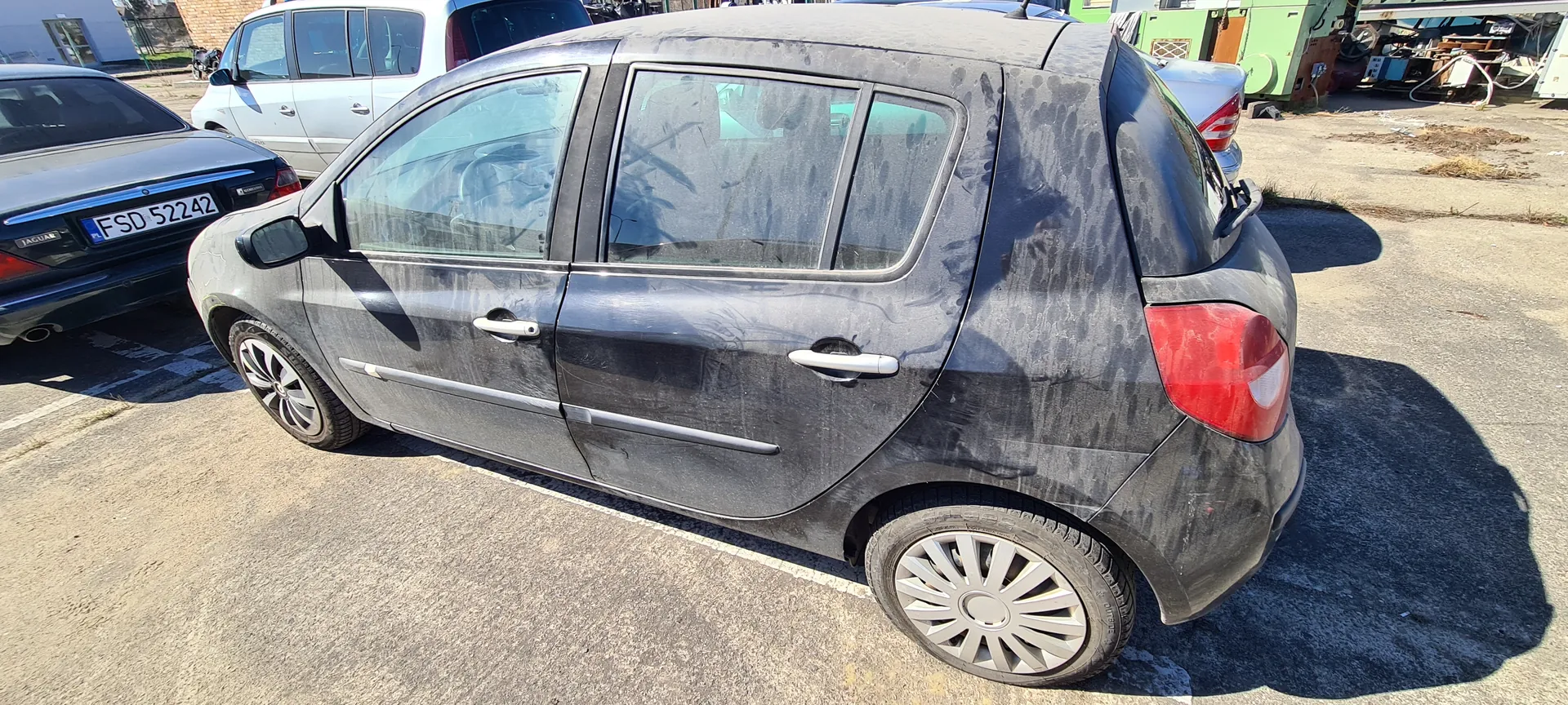 Ciemnoszary samochód osobowy typu hatchback zaparkowany na asfaltowym placu parkingowym. Samochód jest zabrudzony, widoczne są plamy i ślady brudu na karoserii. W tle znajdują się inne pojazdy: czarny samochód z tablicą rejestracyjną FSD 52242 oraz biały van. W oddali widoczne są zielone budynki i różne przedmioty. Słoneczny dzień, cienie wskazują na jasne oświetlenie.