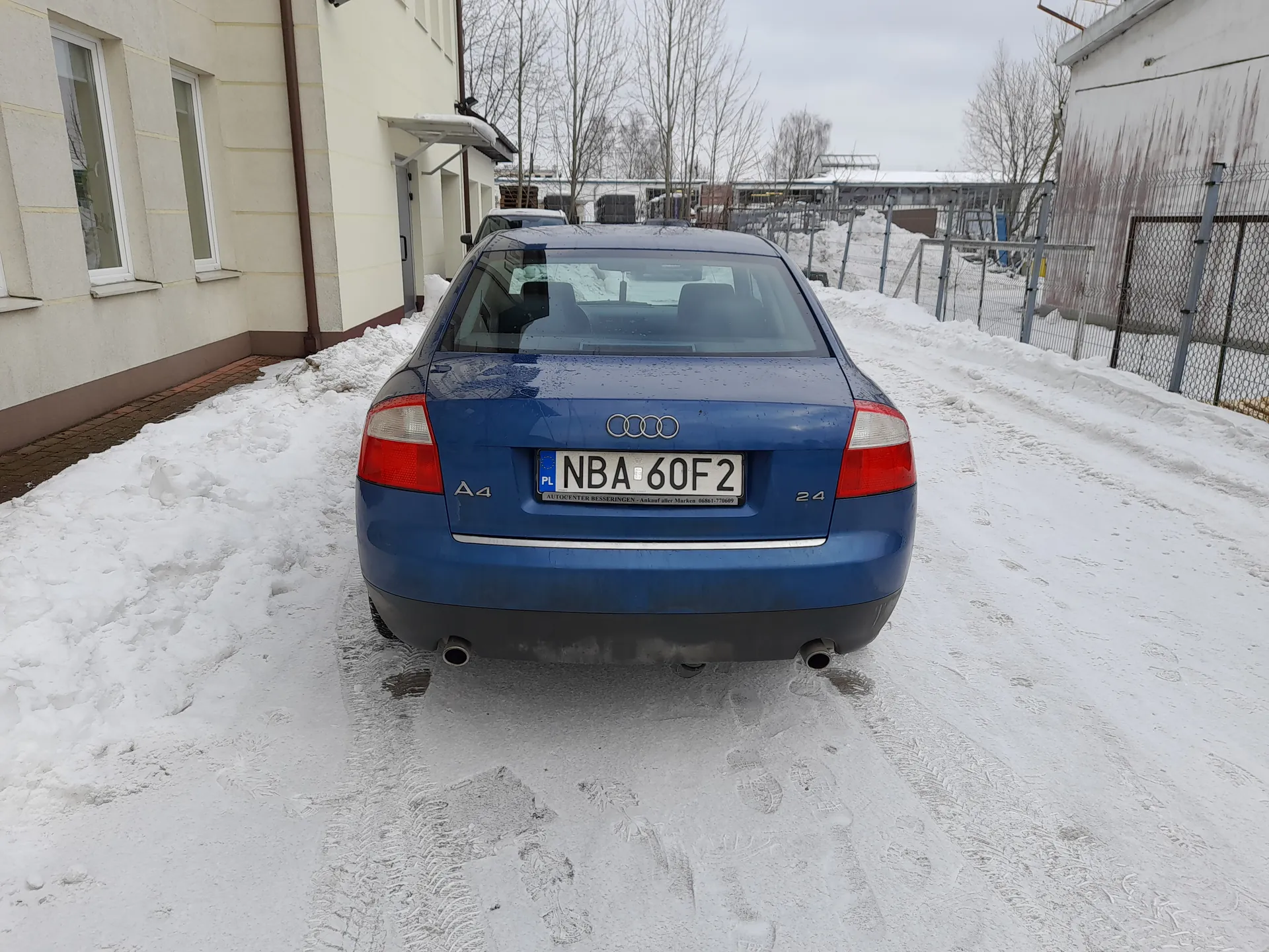 Niebieski samochód Audi A4 z tablicą rejestracyjną PL NBA 60F2 zaparkowany na śnieżnej drodze. Widoczne są budynki po obu stronach, ogrodzenie metalowe po prawej stronie oraz drzewa bez liści na tle. Na tylnym zderzaku widnieją oznaczenia "A4" i "2.4". Droga i obszar wokół są pokryte śniegiem, a na niebie panuje pochmurna pogoda.