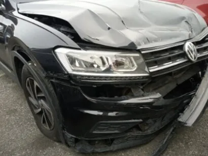 Czarny samochód marki Volkswagen z widocznym logo na atrapie chłodnicy. Przednia część pojazdu jest uszkodzona: zgnieciona maska, pęknięty zderzak i uszkodzony reflektor. Samochód stoi na asfaltowej powierzchni, w tle częściowo widoczny czerwony pojazd.