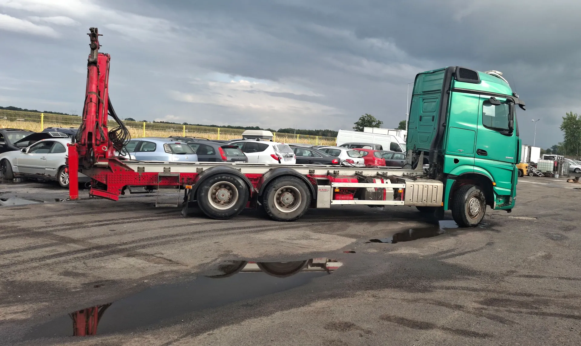 Zielona ciężarówka z czerwonym dźwigiem manipulacyjnym zamontowanym z tyłu platformy, zaparkowana na asfaltowym placu z kałużami. W tle widoczny jest parking z wieloma samochodami osobowych różnych marek i kolorów oraz zachmurzone niebo.