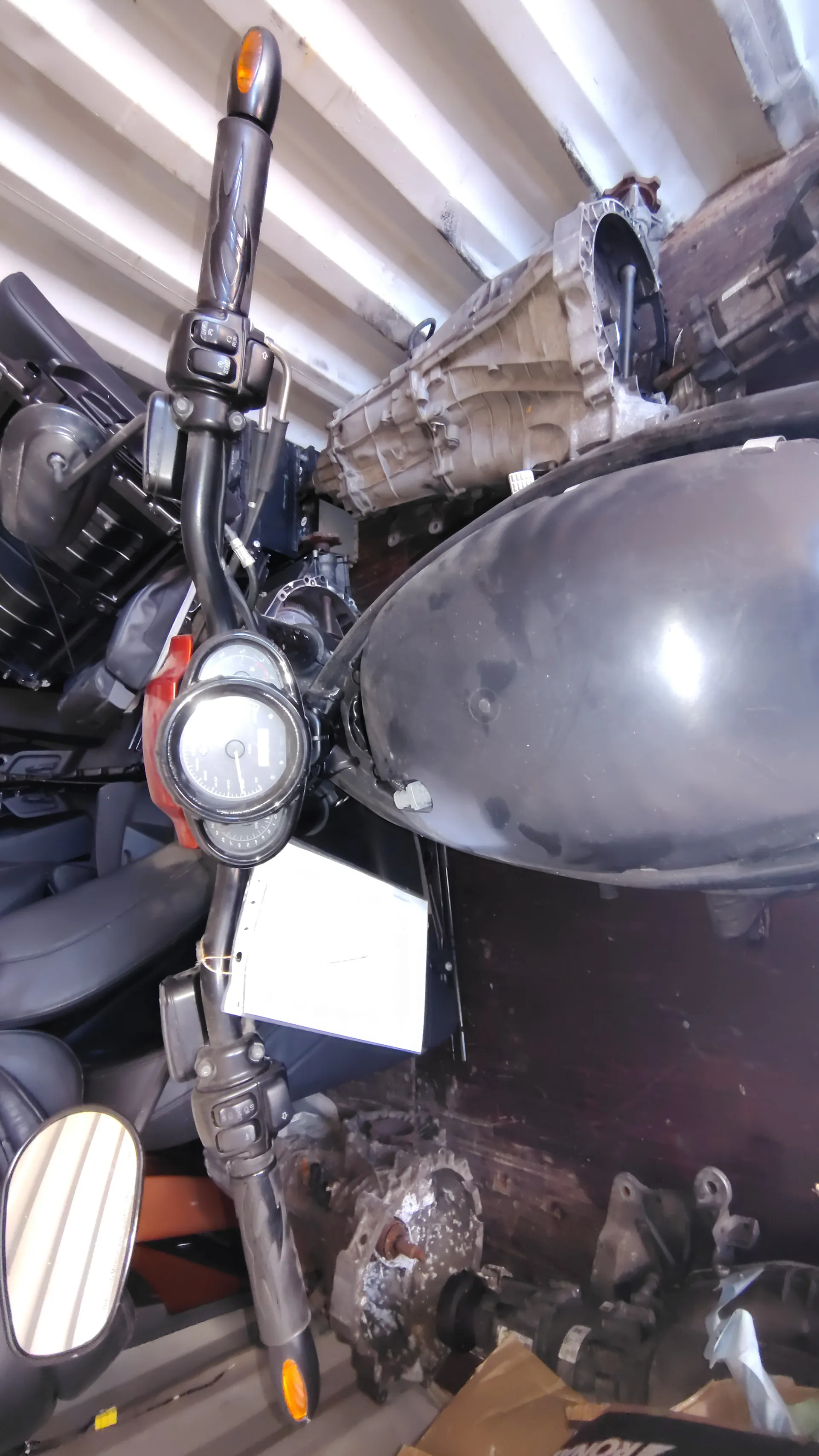 Czarny motocykl z widocznymi kierownicą, licznikami prędkości i białym papierem przypiętym do kierownicy. W tle widoczne są czarne fotele samochodowe oraz różne części mechaniczne wewnątrz kontenera.