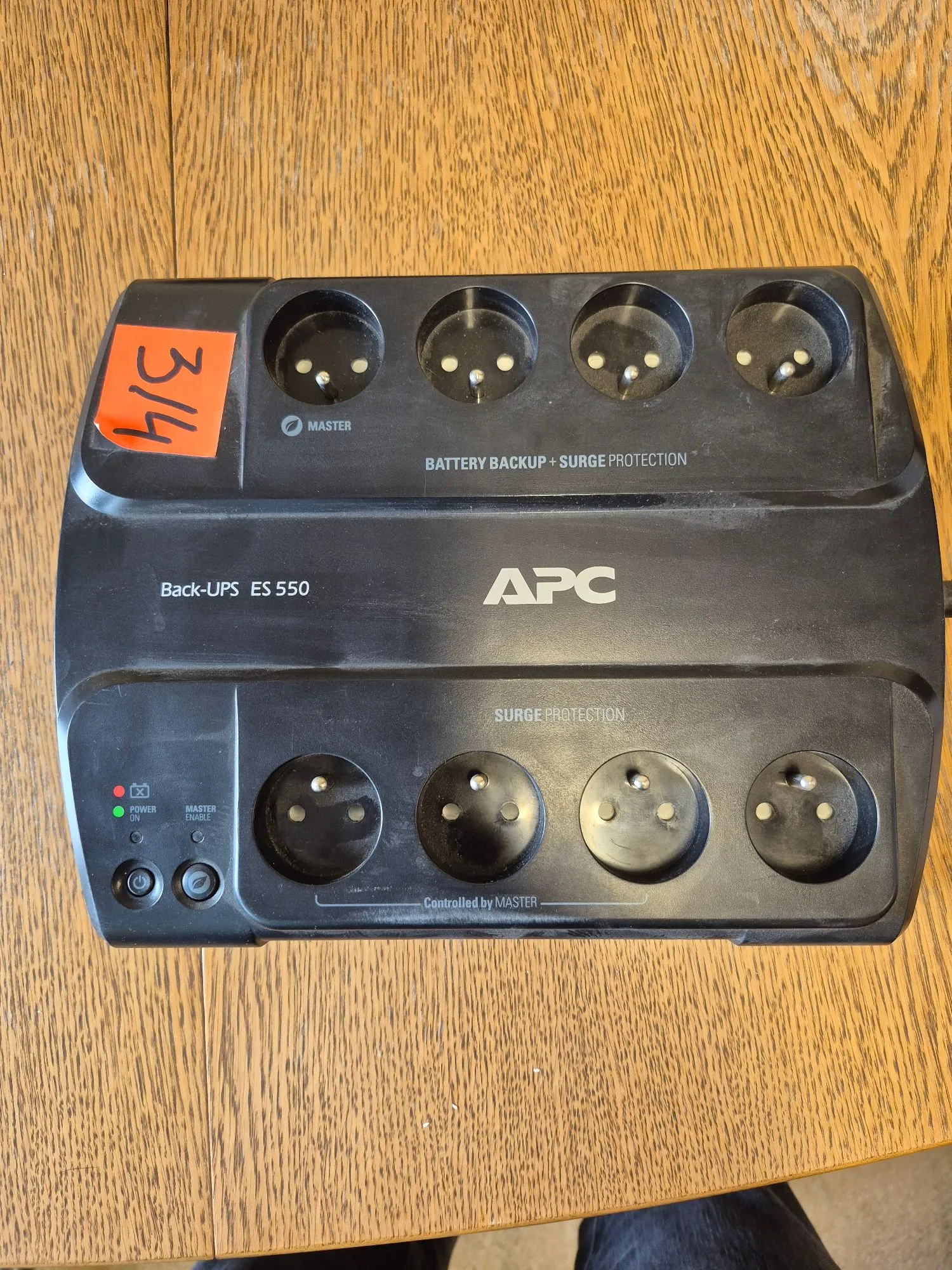Czarny listwa zasilająca marki APC, model Back-UPS ES 550, leżąca na drewnianym blacie. Urządzenie posiada osiem gniazd elektrycznych typu Schuko, podzielonych na dwa rzędy po cztery. Górny rząd zawiera gniazdo oznaczone jako 'MASTER' oraz napis 'BATTERY BACKUP + SURGE PROTECTION'. Dolny rząd zawiera gniazda oznaczone jako 'Controlled by MASTER' oraz napis 'SURGE PROTECTION'. Po lewej stronie znajdują się dwa przyciski: 'POWER' z diodą oraz 'MASTER ENABLE'. W lewym górnym rogu naklejona jest pomarańczowa etykieta z ręcznie napisanym numerem '314'.