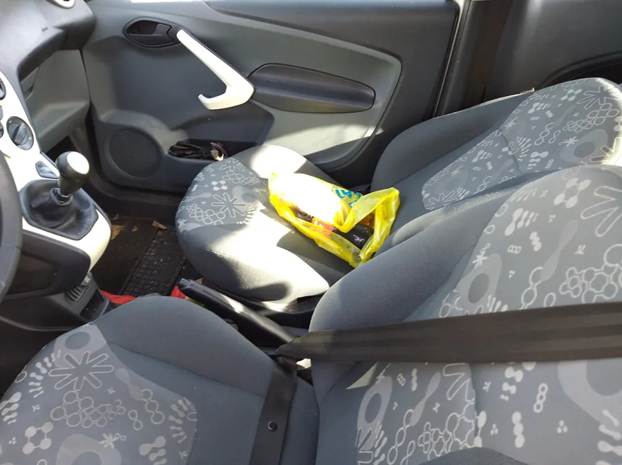 Wnętrze samochodu z widocznymi przednimi siedzeniami w szaro-białym wzorze. Na pasażerskim siedzeniu leży żółta torba z plastiku. Po lewej stronie widoczna jest kierownica, dźwignia zmiany biegów oraz fragment deski rozdzielczej. Drzwi pasażerskie z uchwytem i głośnikiem. Na podłodze widoczny dywanik i fragment czarnego przedmiotu w pobliżu drzwi. Pas bezpieczeństwa przewieszony przez siedzenie pasażera.