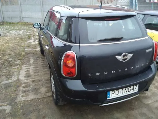 Czarny samochód Mini Countryman widziany od tyłu, z widocznym logo MINI z napisem COUNTRYMAN na bagażniku. Tablica rejestracyjna PO 1NG47 umieszczona na dole. Samochód zaparkowany na betonowej powierzchni z plamami trawy, w tle szara ściana i częściowo widoczny żółty samochód po prawej stronie.