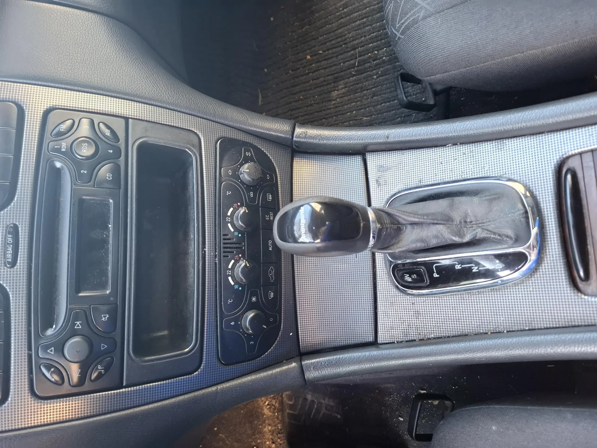 Wnętrze samochodu, widok na centralną konsolę z dźwignią zmiany biegów oznaczoną literami P, R, N, D, 2, 1. Po lewej stronie panel klimatyzacji z pokrętłami i przyciskami, w tym oznaczeniami AUTO, EC, REST oraz skalą temperatury. Obok znajduje się radio z przyciskami (1, 2, 3, 4, CD, RDS) i wyświetlaczem. Konsola wykończona teksturą podobną do metalu, siedzenia tapicerowane szarym materiałem. Widoczny napis "AIRBAG OFF" po lewej stronie.