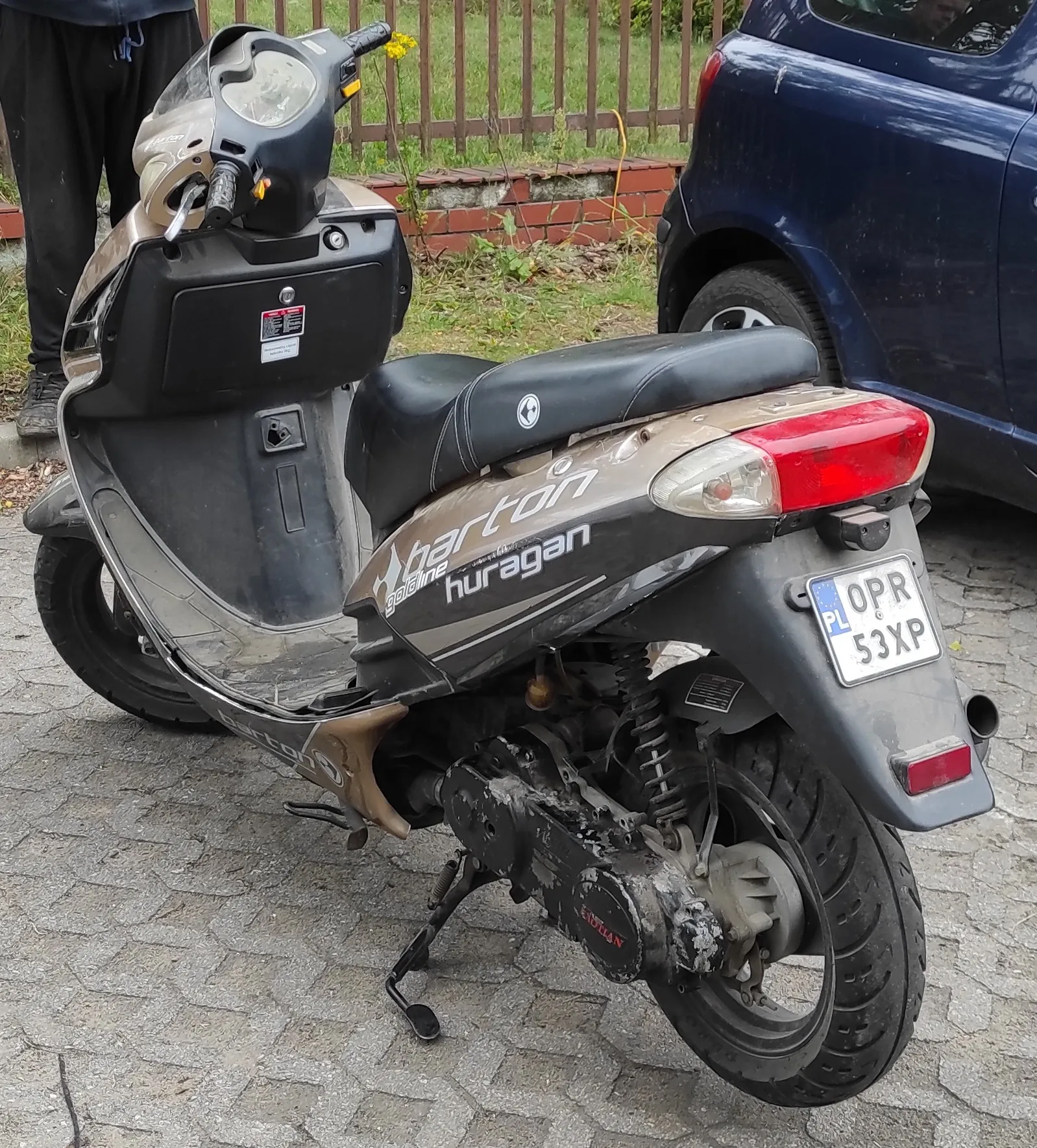 Widok z tyłu i z boku brązowo-czarnego skutera marki Barton, model Huragan. Skuter posiada polską tablicę rejestracyjną o numerze OPR 53XP. Pojazd stoi zaparkowany na kostce brukowej obok ciemnoniebieskiego samochodu i drewnianego ogrodzenia.