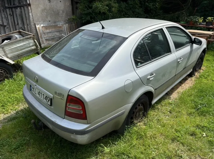 Srebrny samochód osobowy marki Skoda Octavia zaparkowany na trawie. Widok z tyłu i z boku. Pojazd posiada polską tablicę rejestracyjną o numerze ZKL 41149. W tle widoczny jest drewniany płot, fragment przyczepy oraz zieleń ogrodowa.