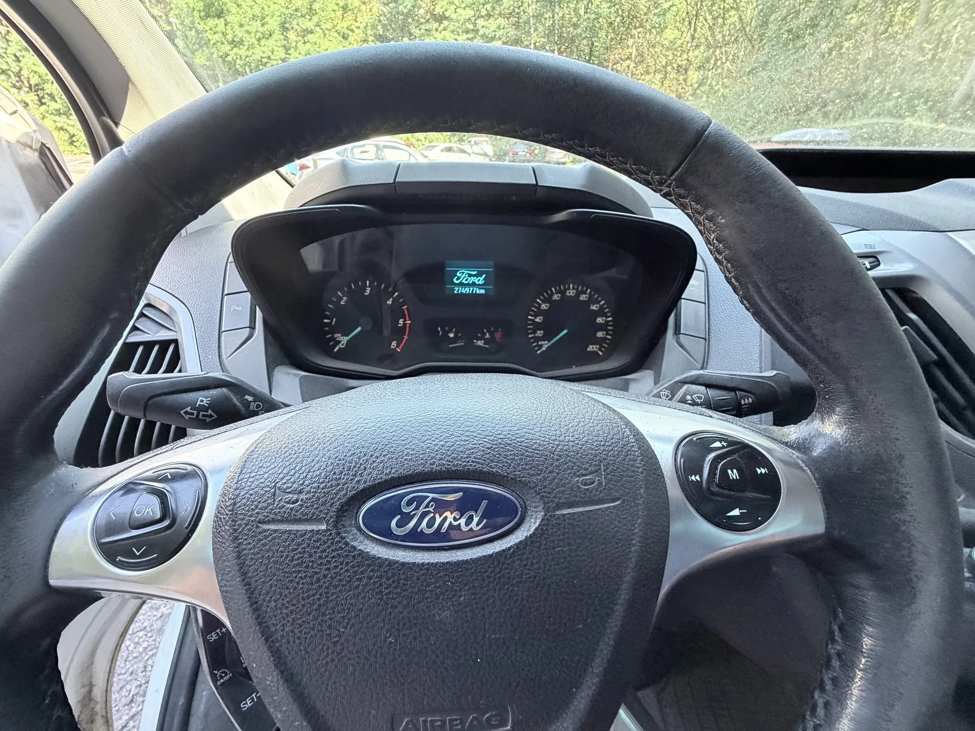 Widok z perspektywy kierowcy na wnętrze samochodu marki Ford. Na pierwszym planie znajduje się czarna, skórzana kierownica z logo Ford na środku oraz przyciskami sterującymi po bokach. W tle widoczny jest panel instrumentów z prędkościomierzem, obrotomierzem i wyświetlaczem cyfrowym wskazującym przebieg 274977 km. Przez przednią szybę widać zielone drzewa.