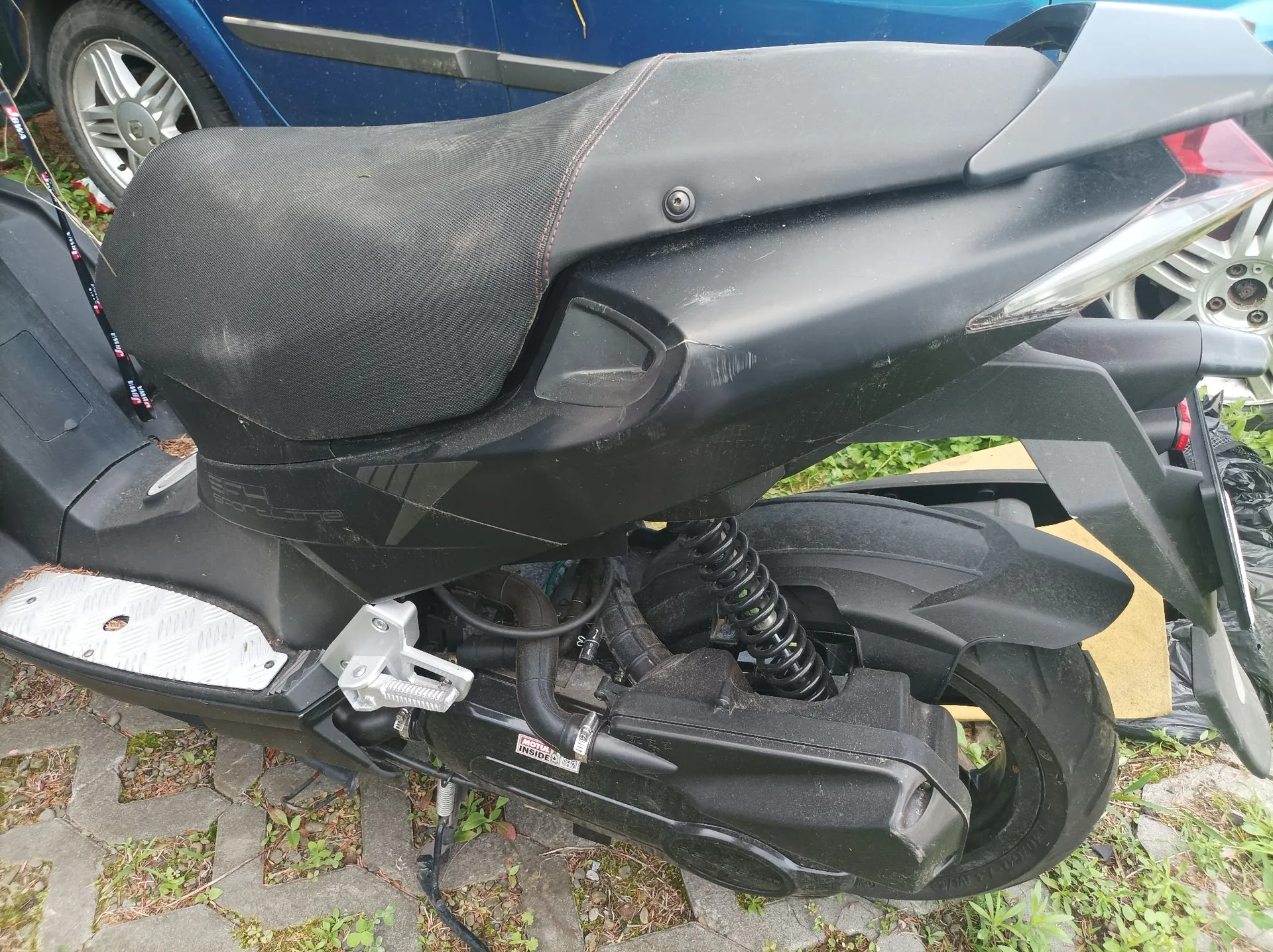 Zbliżenie na tylną część czarnego skutera zaparkowanego na kostce brukowej z trawą. Widoczne jest czarne, teksturowane siedzenie, tylny amortyzator z czarną sprężyną oraz tylne koło. W tle znajduje się fragment niebieskiego samochodu.