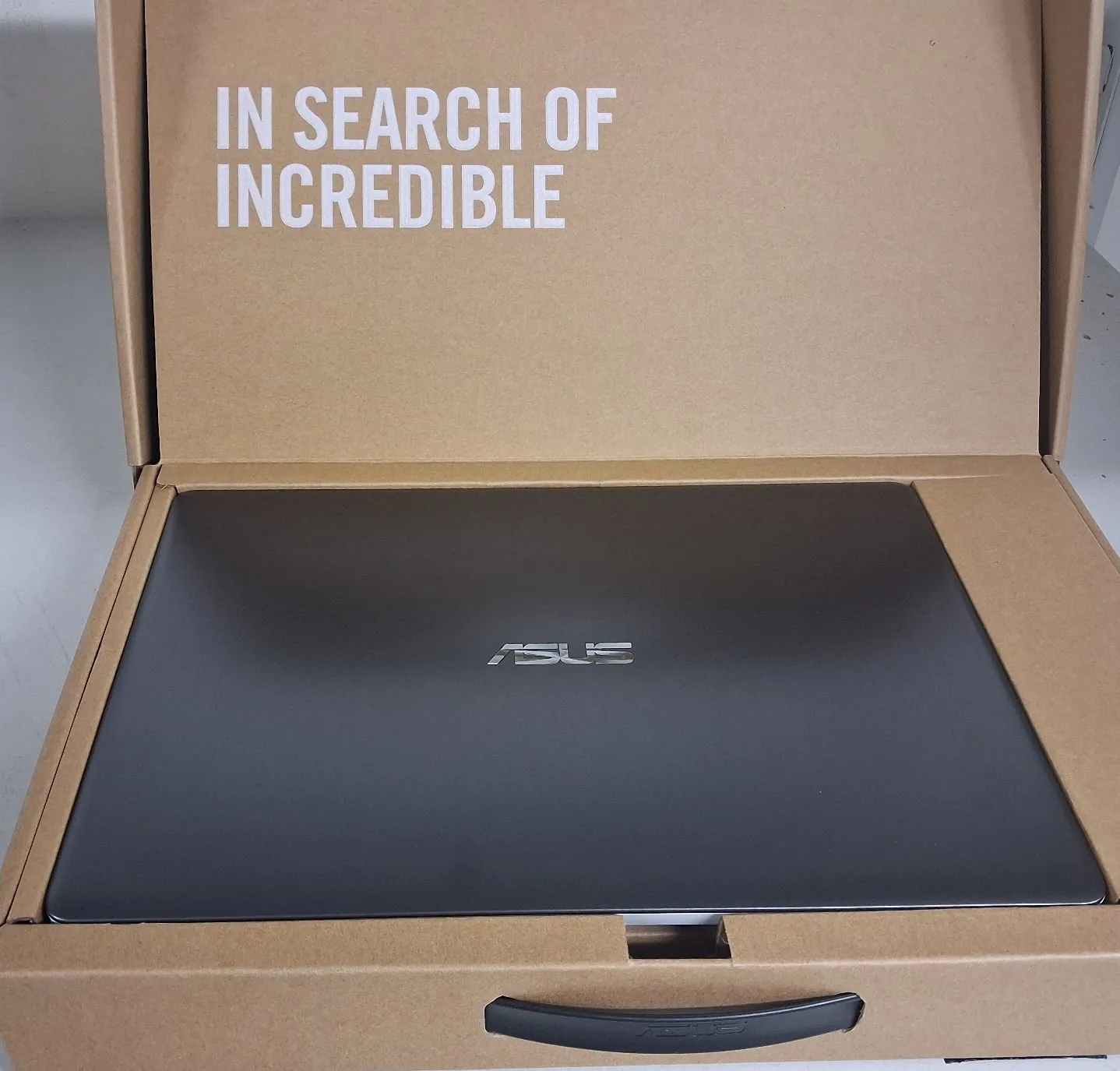 Otwarta brązowa kartonowa коробka, w której znajduje się czarny laptop marki ASUS. Na wewnętrznej stronie klapy pudełka widnieje biały napis: 'IN SEARCH OF INCREDIBLE'.