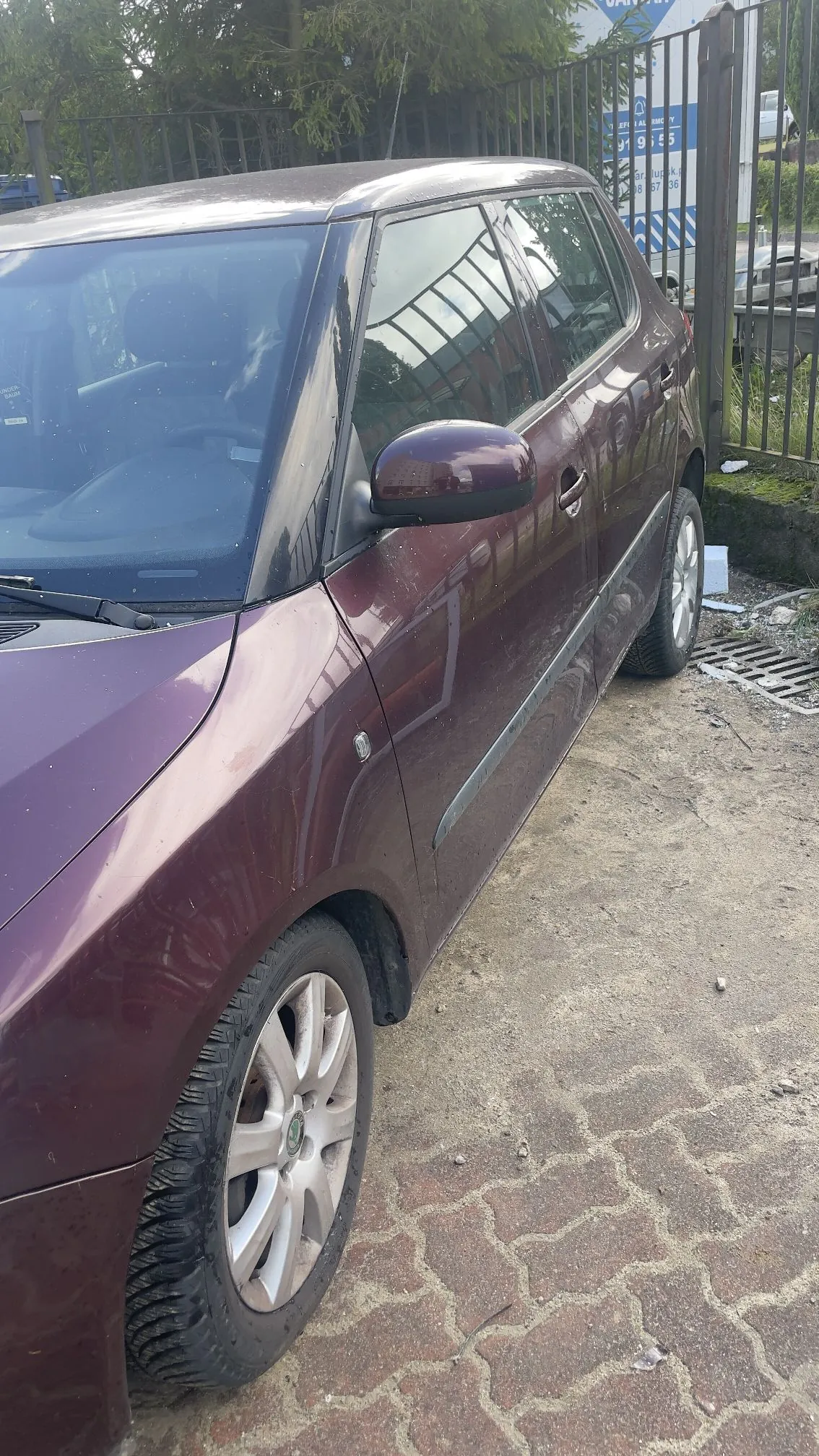 Boczny widok bordowego samochodu osobowego zaparkowanego na kostce brukowej obok metalowego ogrodzenia. Widoczna jest przednia i tylna prawa strona pojazdu, lusterko boczne oraz felgi aluminiowe.